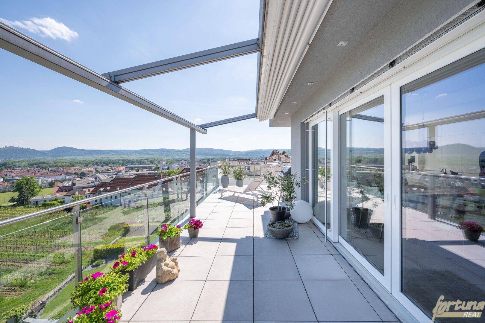 Große Dachterrasse mit Glasgeländer, Fliesenboden und weitem Blick über die Landschaft und Weinberge.