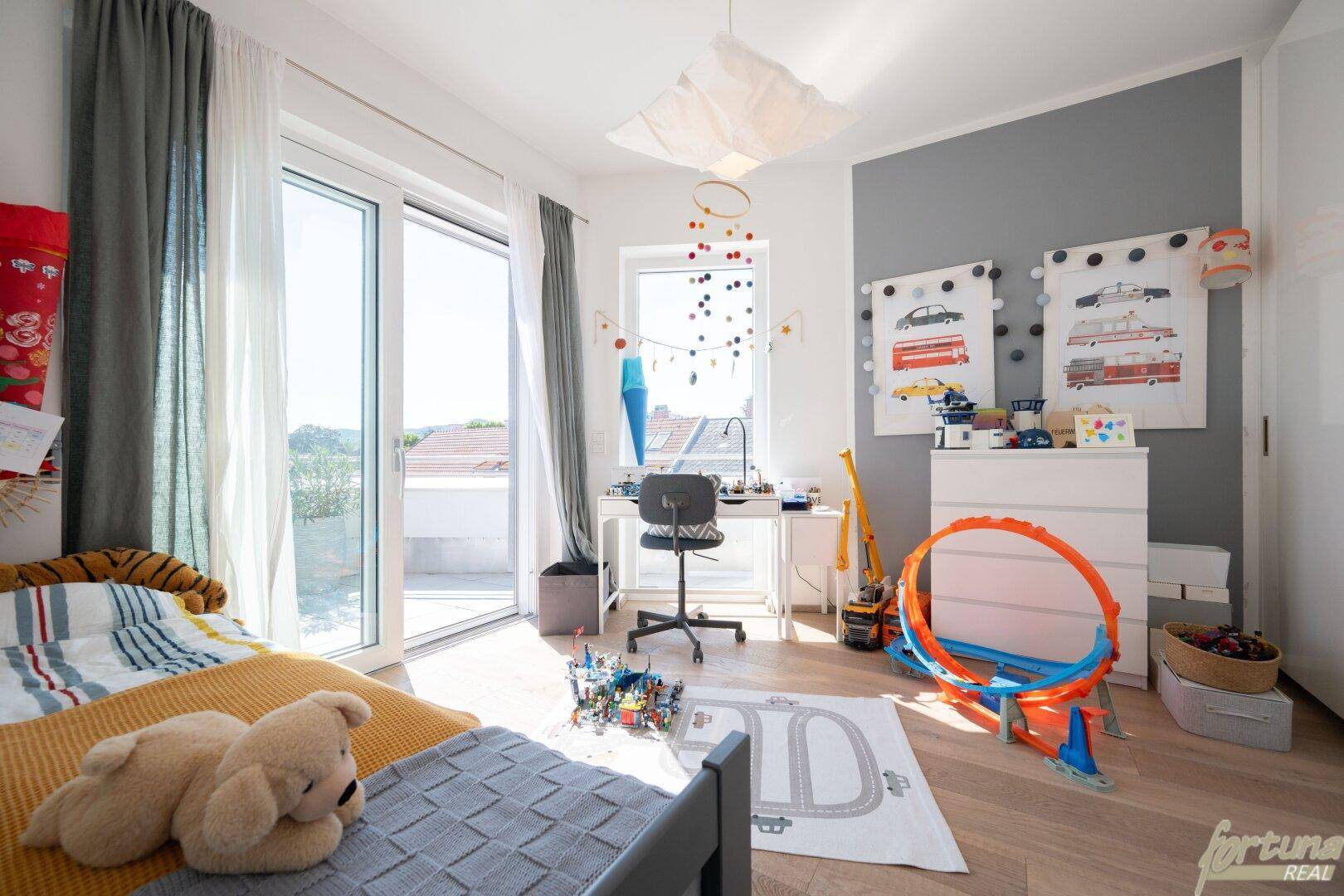 Kinderzimmer mit Holzboden, Schreibtisch, Spielzeug und großen Fenstern mit Zugang zum Außenbereich.