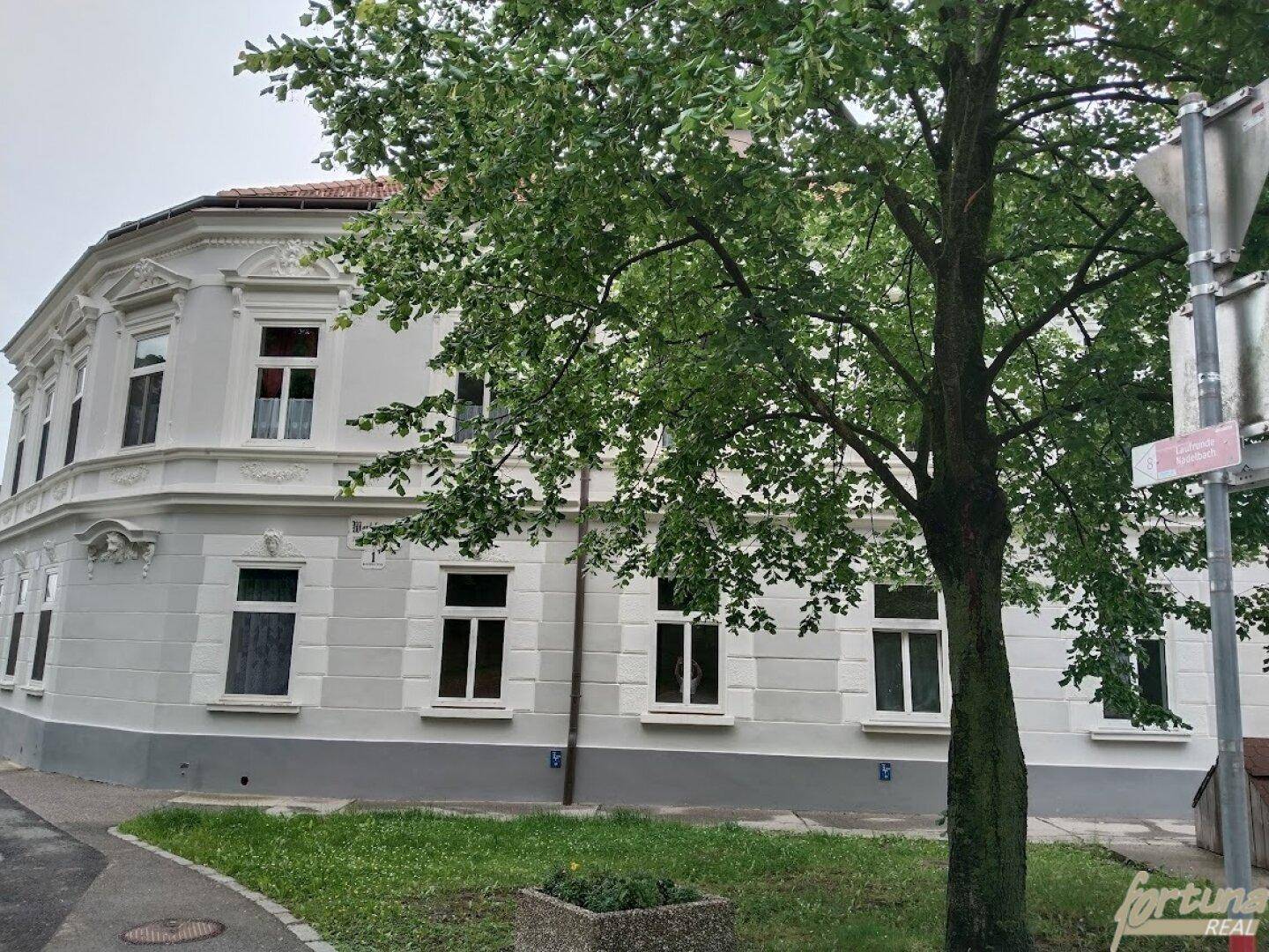 Historisches Gebäude mit klassischer Fassade und großen Fenstern, umgeben von Grünflächen und Bäumen.