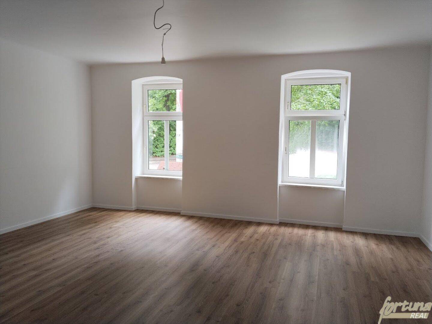 Geräumiges Wohnzimmer mit Laminatboden und zwei großen Fenstern, die einen Blick ins Grüne bieten.