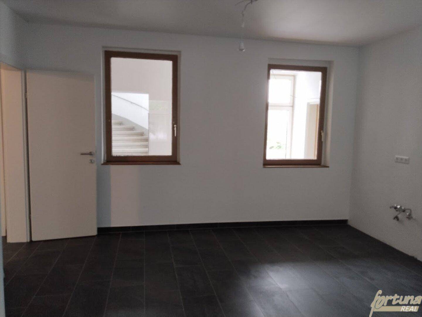 Leerer Raum mit dunklen Fliesen und Fenstern, die einen Blick auf das Treppenhaus ermöglichen.