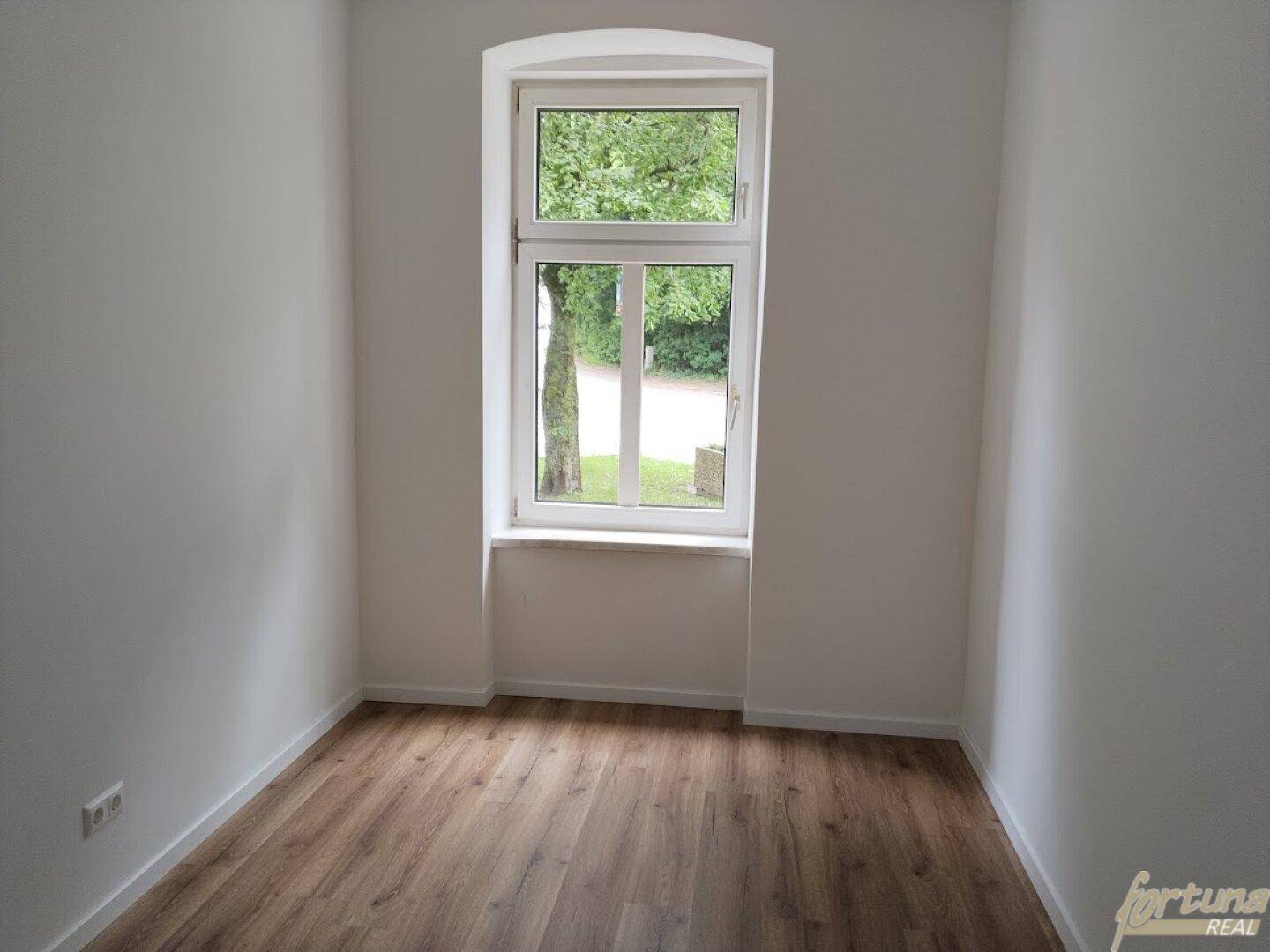 Kleines, helles Zimmer mit Laminatboden und einem Fenster, das viel Tageslicht hereinlässt und Bäume zeigt.