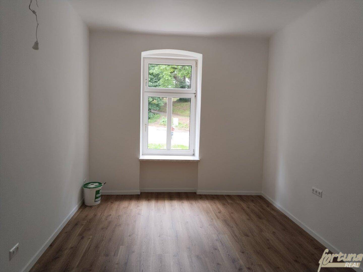 Helles Zimmer mit Laminatboden und einem großen Fenster, das viel Tageslicht hereinlässt und eine grüne Aussicht bietet.