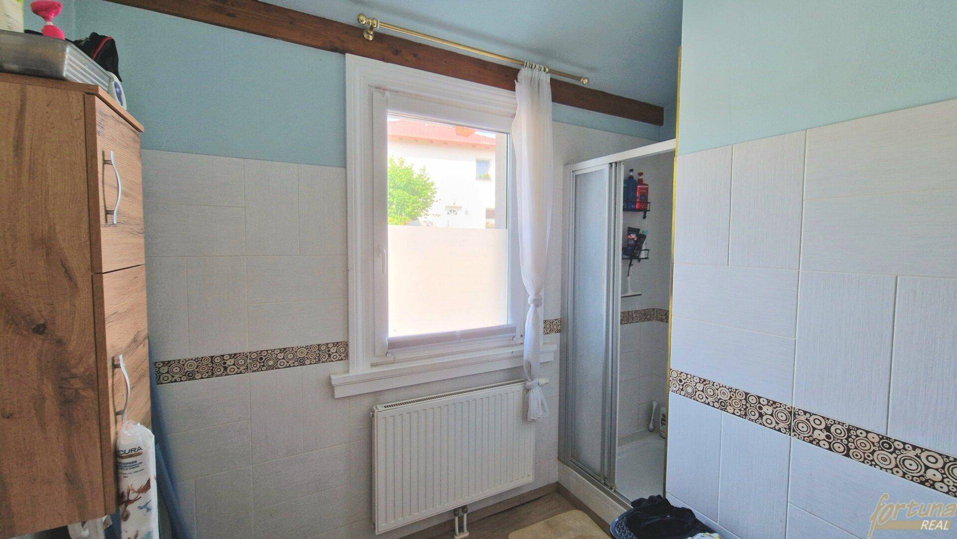 Badezimmer mit Duschkabine, Fenster und hellen Fliesen, teilweise mit Dekorstreifen.