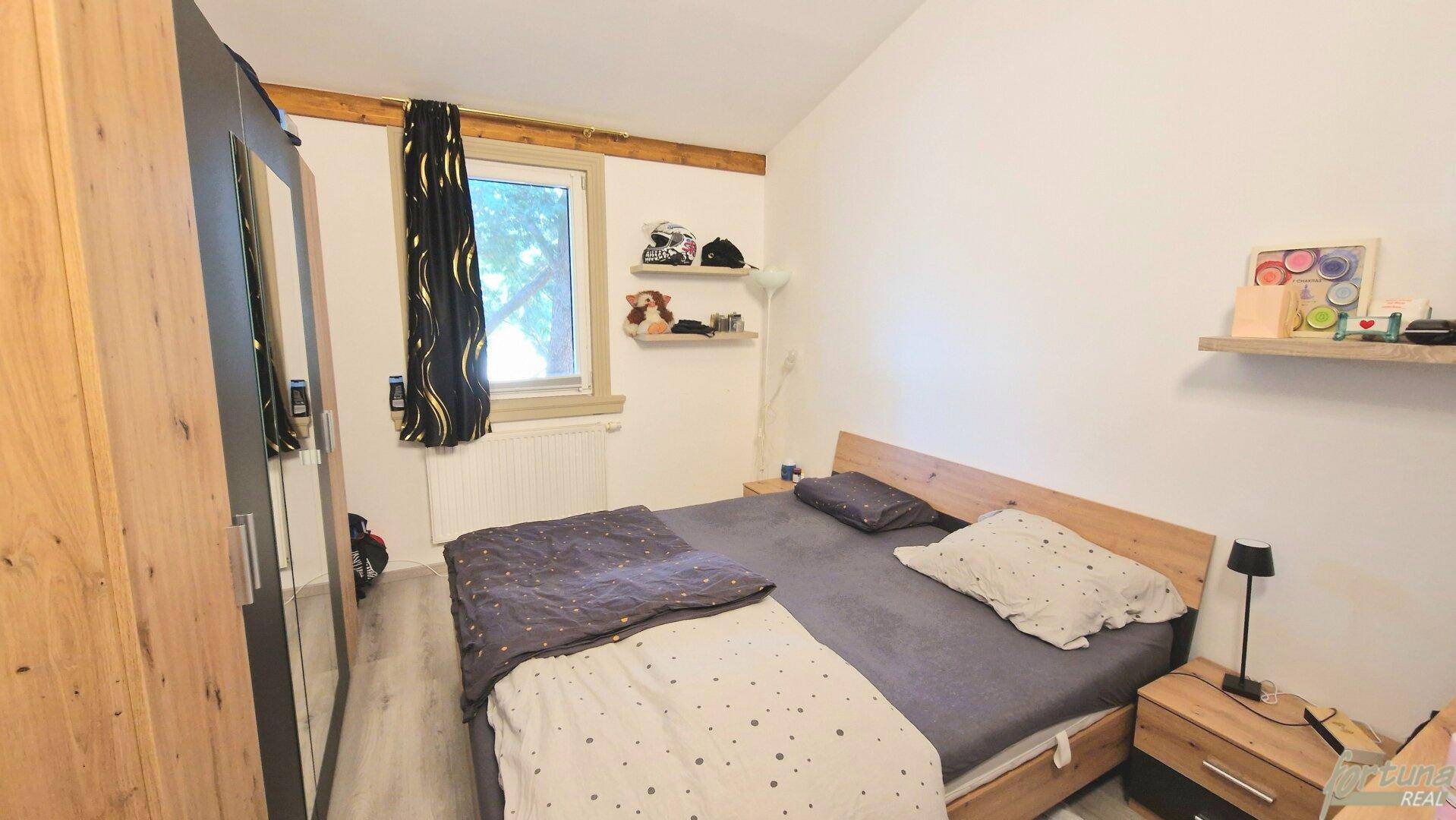 Schlafzimmer mit Doppelbett, Holzschrank und Fenster, das viel Tageslicht hereinlässt.