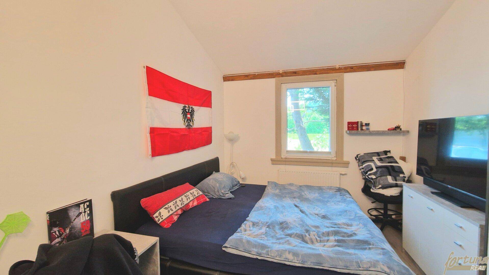 Schlafzimmer mit Bett, Fernseher und einem Fenster, das den Blick ins Grüne freigibt.