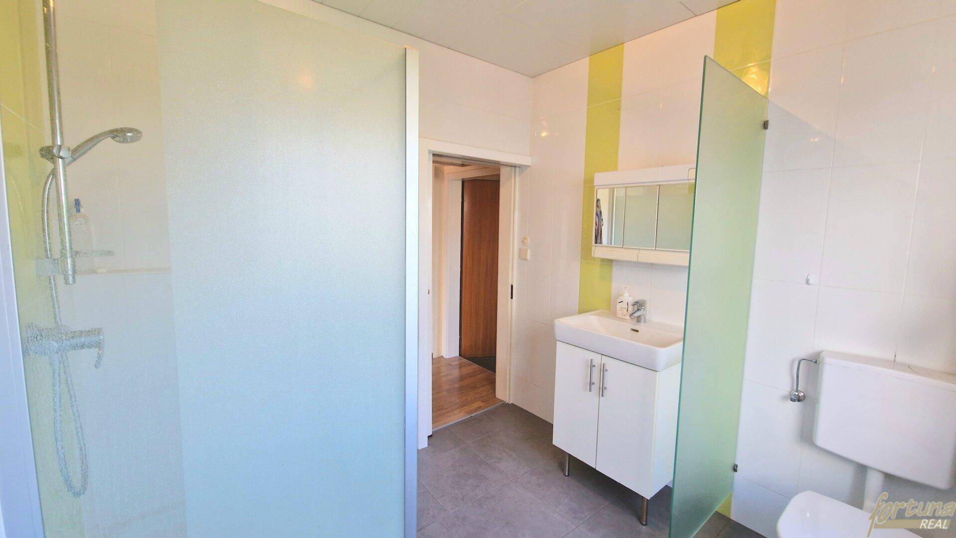 Modernisiertes Badezimmer mit Dusche, Waschbecken und WC, gefliest in Gelb-Weiß, gut gepflegt.
