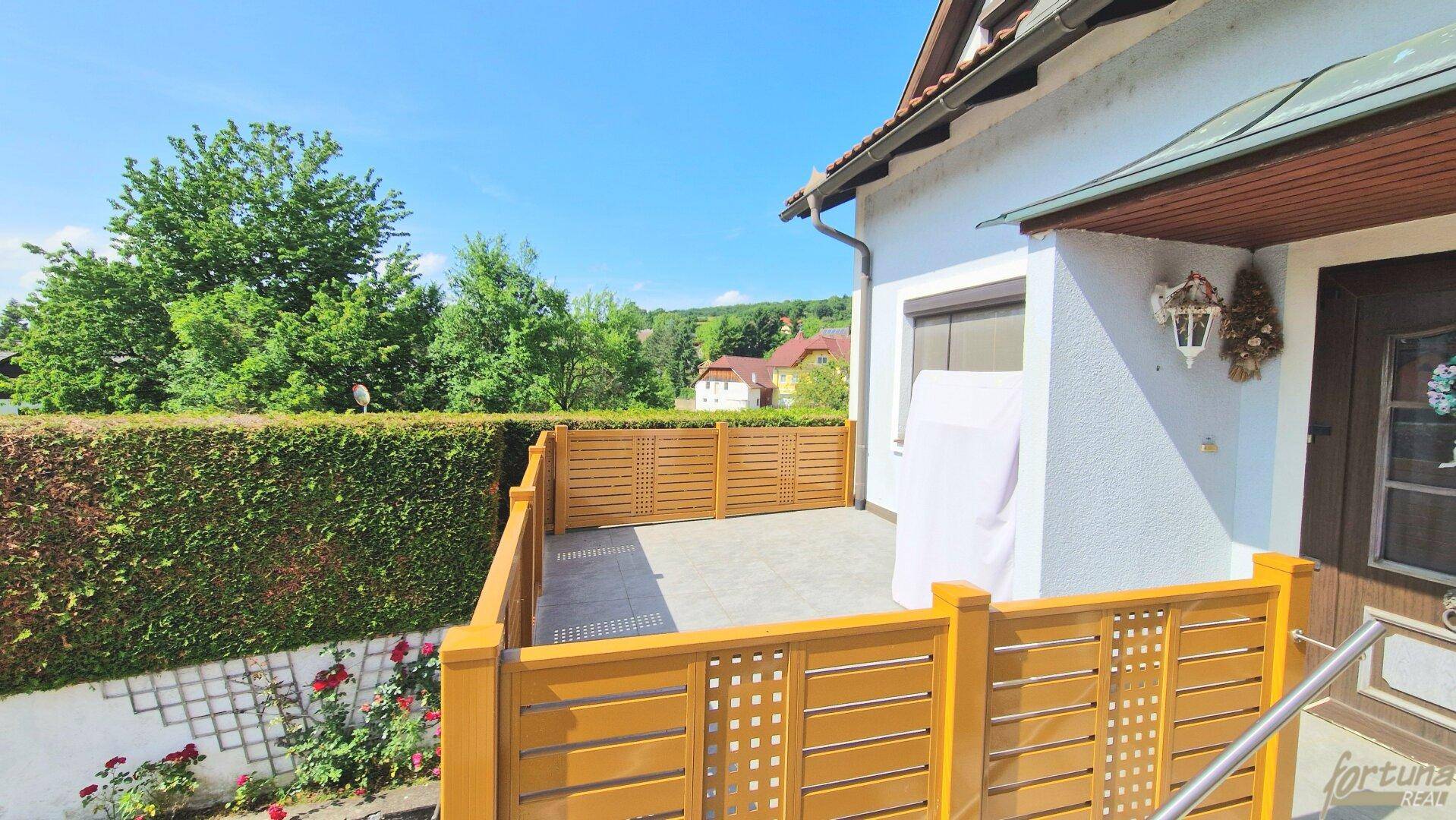 Sonniger Terrassenbereich mit Holzzaun, gepflastertem Boden und grüner Hecke, ideal zum Entspannen.