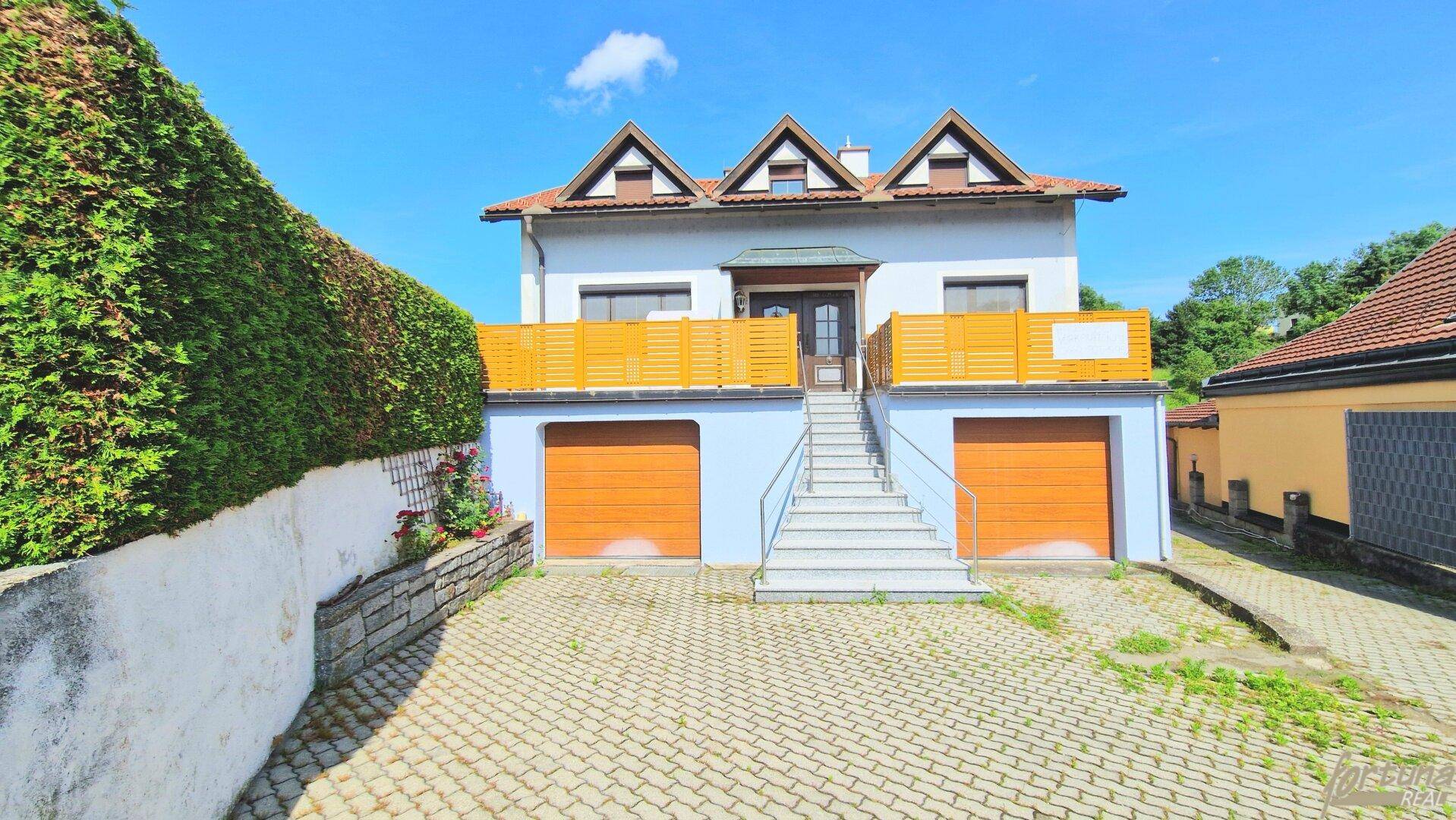 Hausfassade mit großer Terrasse über den Garagen und Außentreppe unter blauem Himmel.