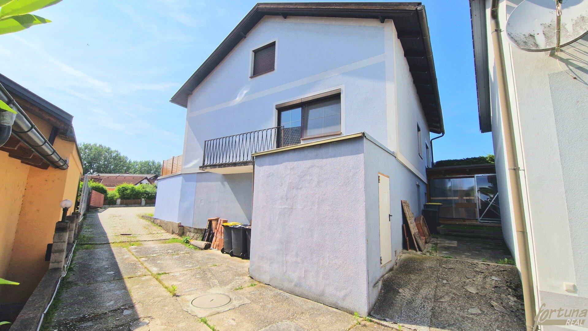 Seitenansicht des Hauses mit schmaler Zufahrt und kleinem Balkon, umgeben von Grün.