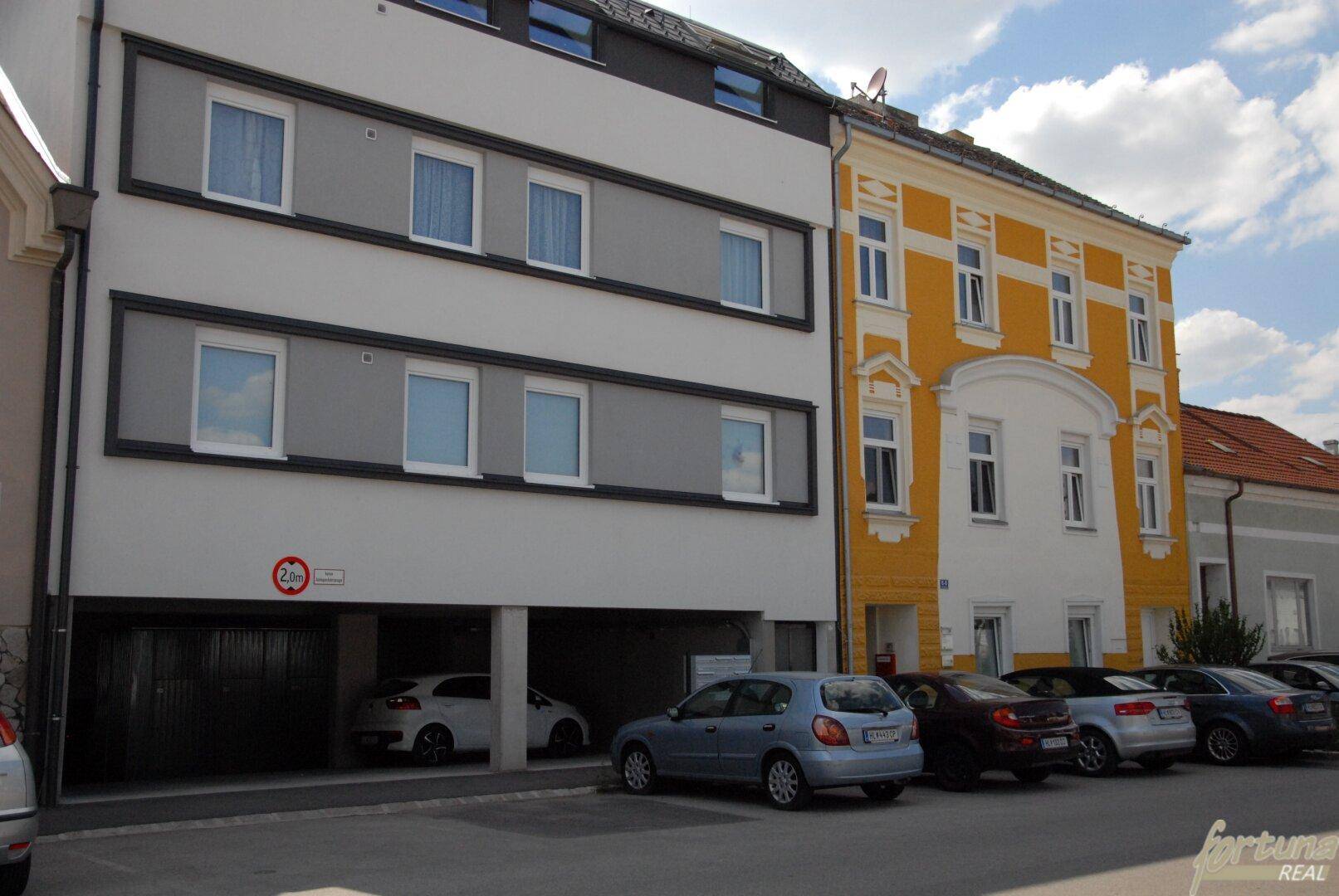 Gebäudeansicht mit moderner weißer Fassade, Tiefgarageneinfahrt und Außenparkplätzen.