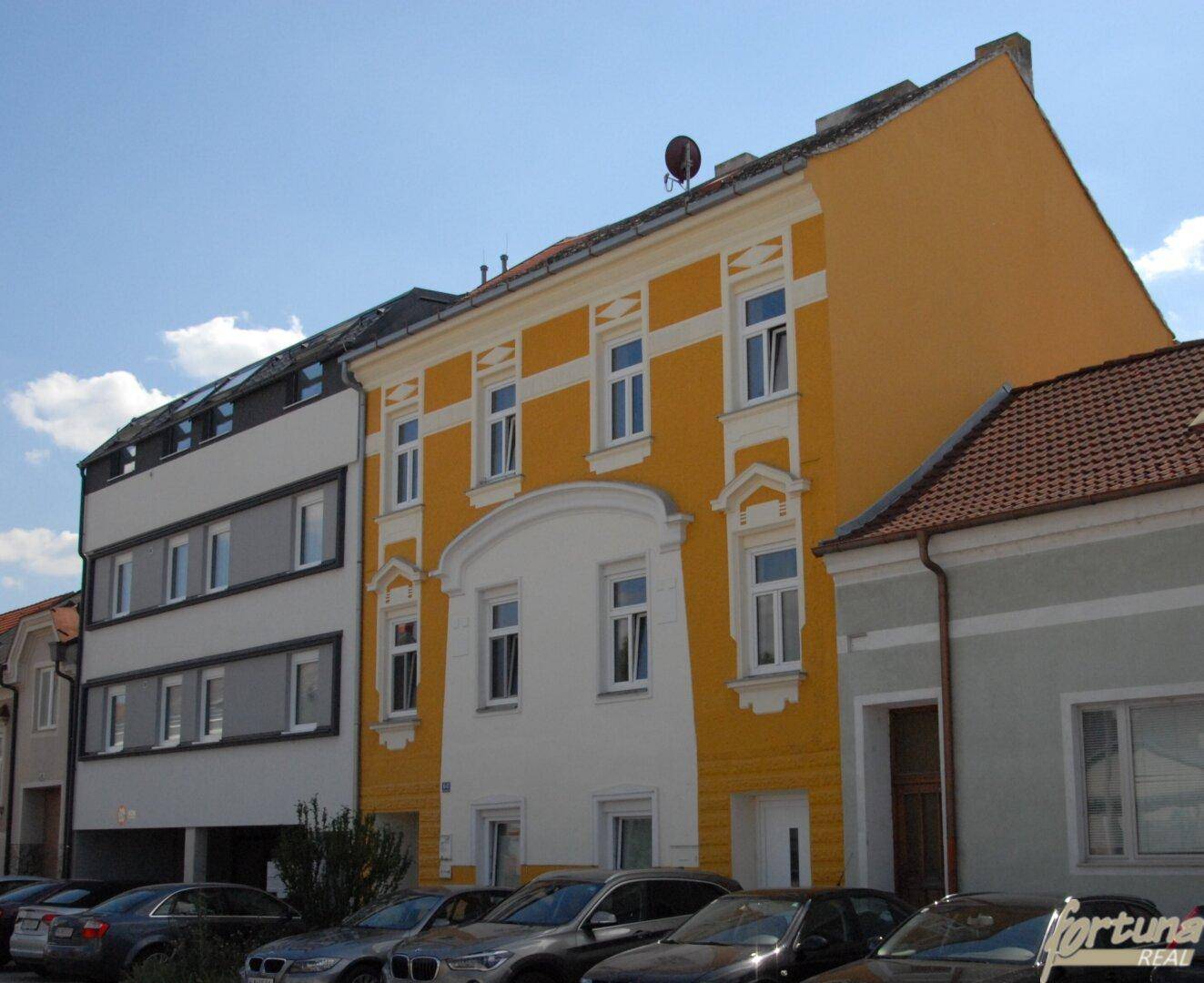 Ansicht eines Gebäudes mit gelber und weißer Fassade sowie parkenden Autos davor.