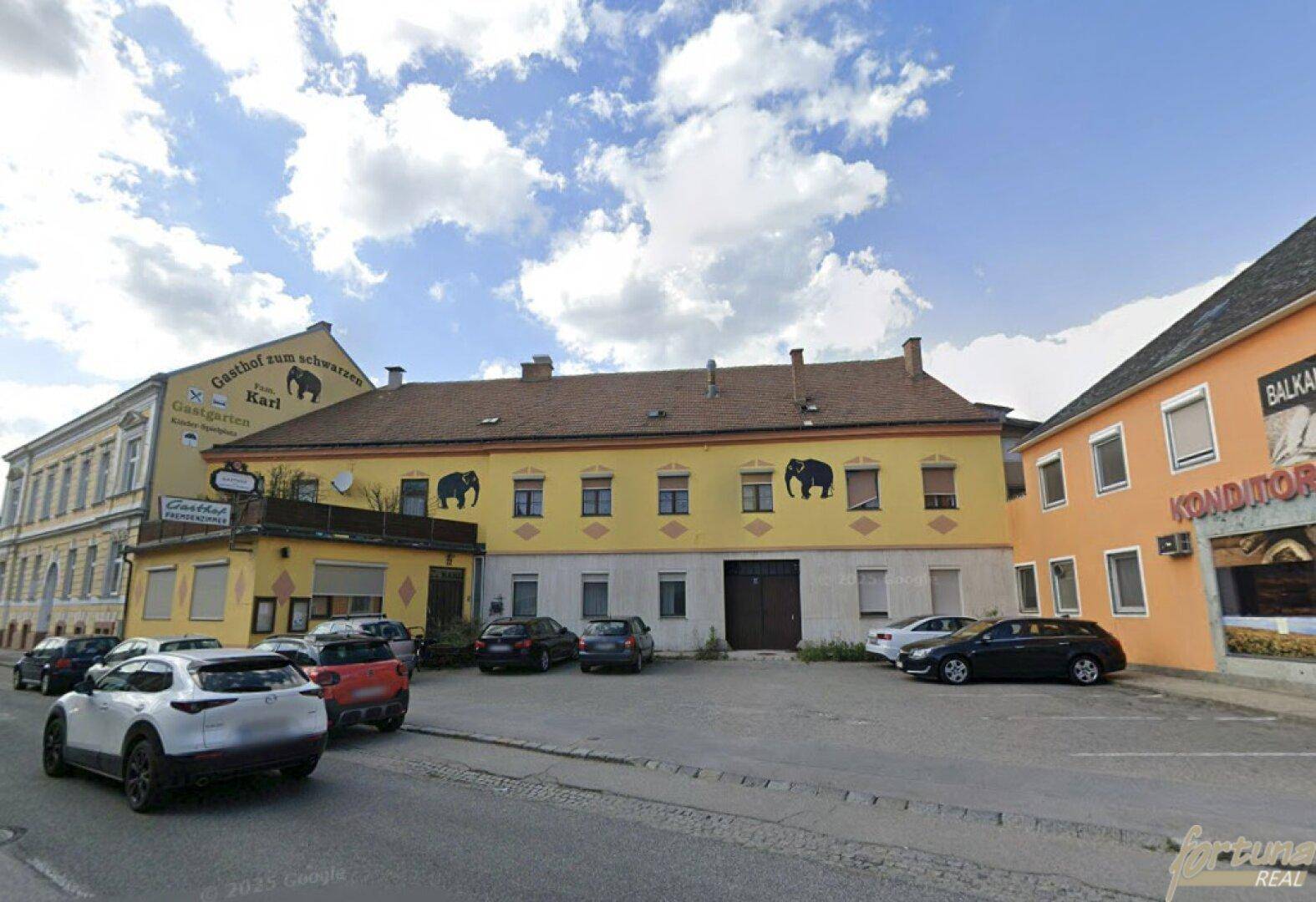 Gelbes Gebäude mit Elefantenmalereien, einem Gasthof-Schild und parkenden Autos davor.
