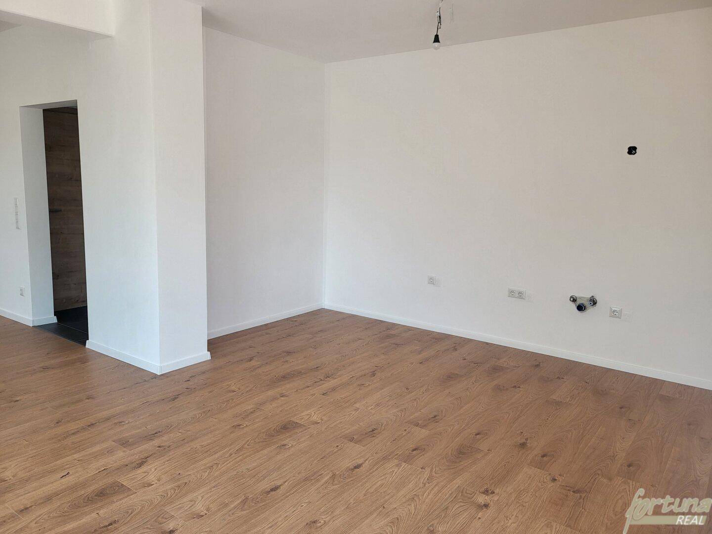 Leerer Küchenbereich mit Holzboden und vorbereiteten Anschlüssen für eine Einbauküche.