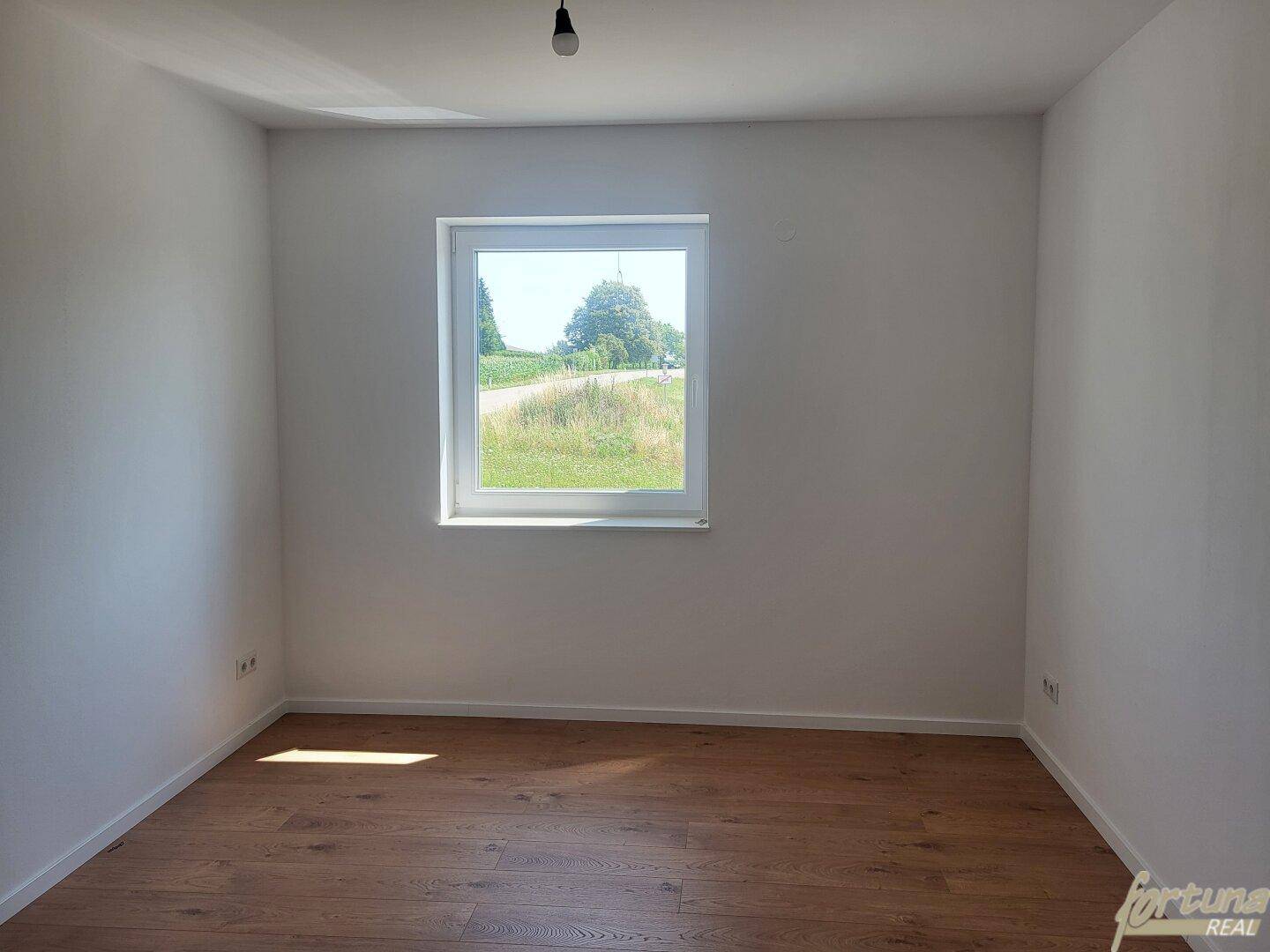 Helles Zimmer mit Holzboden und einem Fenster, das den Blick ins Grüne freigibt.
