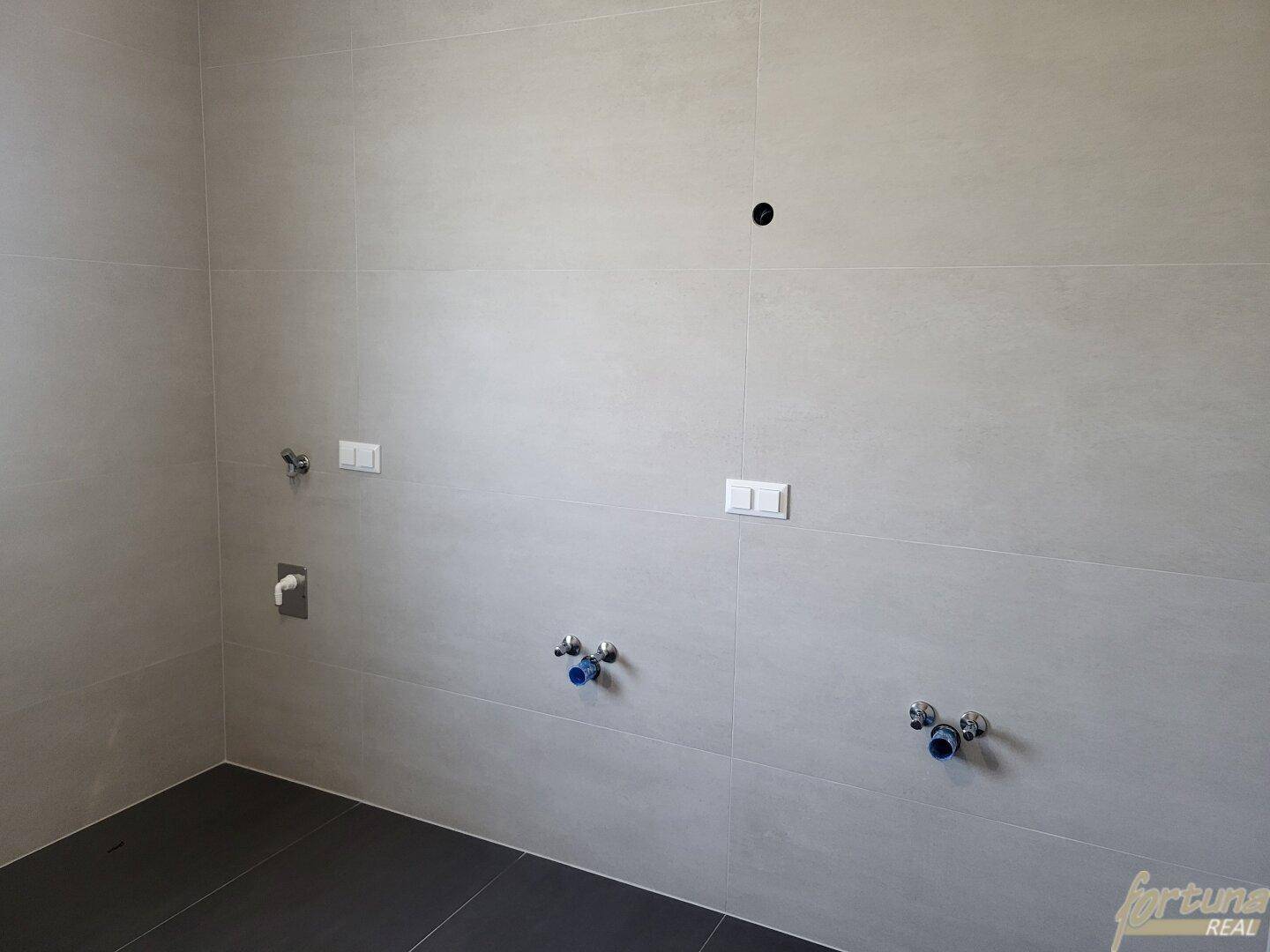 Badezimmer im Rohbau mit hellgrauen Fliesen und vorbereiteten Anschlüssen für Sanitärobjekte.