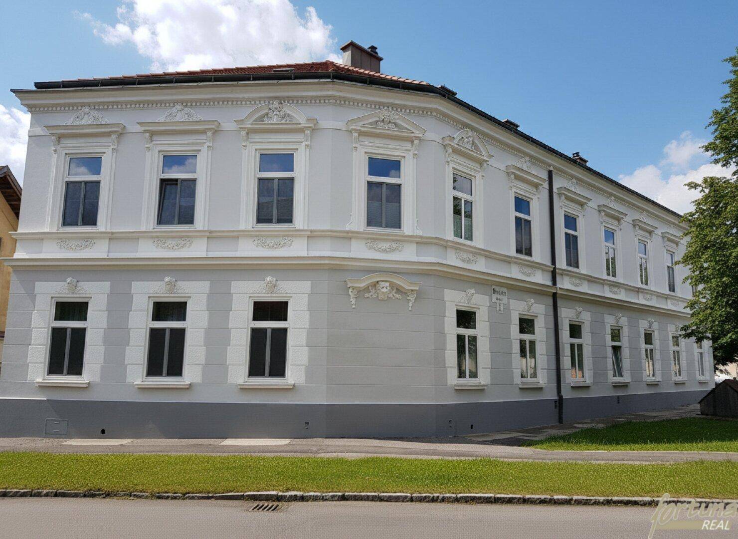 Elegantes Eckgebäude mit klassischer Architektur und großen Fenstern, umgeben von Grünflächen unter blauem Himmel.