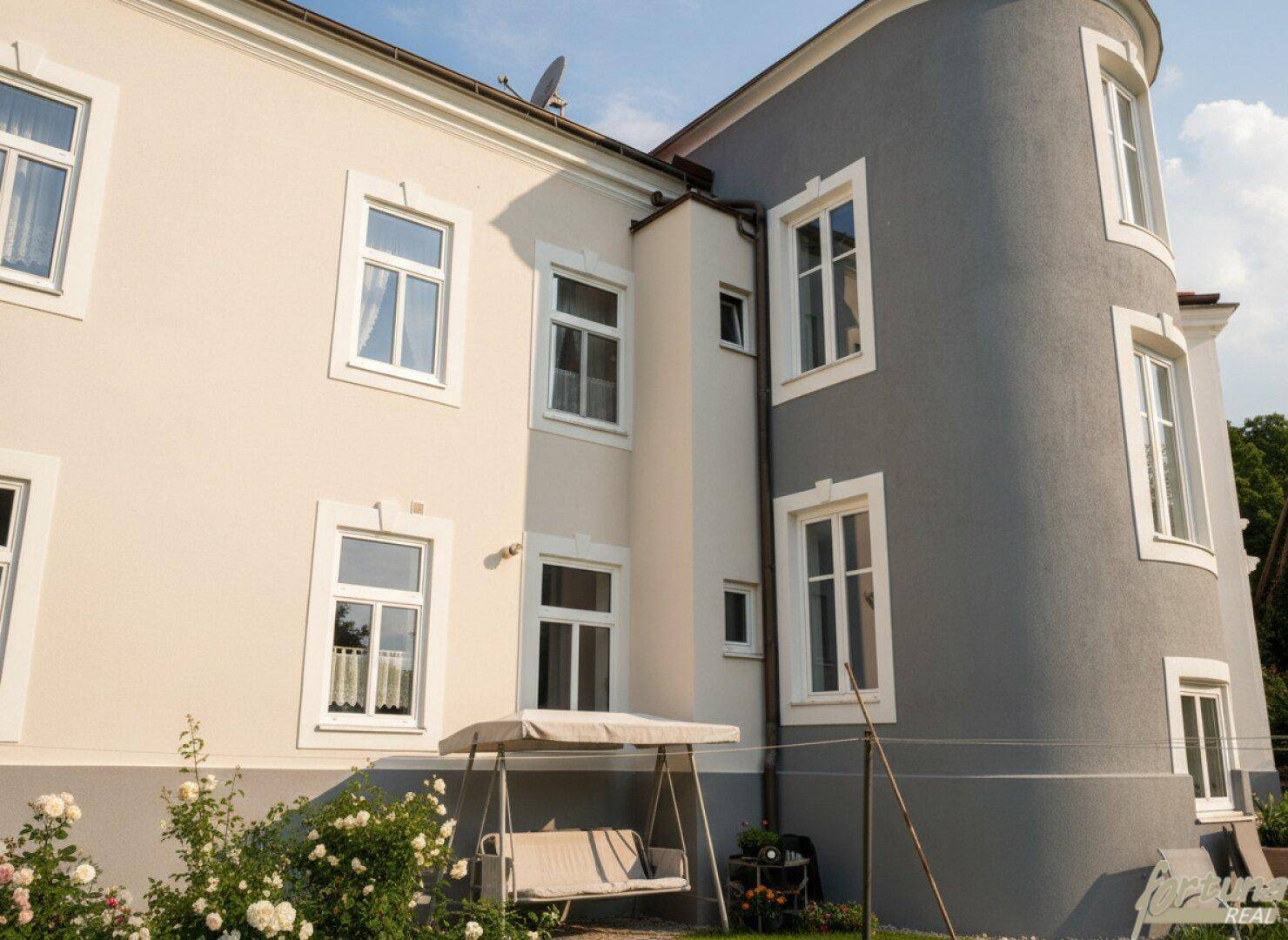 Ruhiger Gartenbereich an der Gebäudeseite mit einer Schaukel und blühenden Sträuchern unter blauem Himmel.