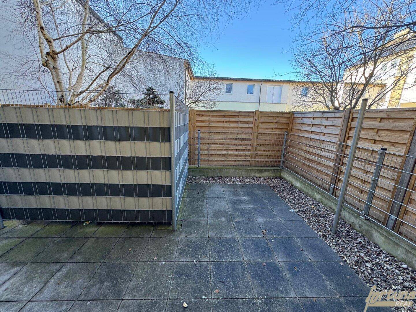 Kleine, gepflasterte Terrasse mit Holzzaun und Sichtschutz, umgeben von kahlen Bäumen.