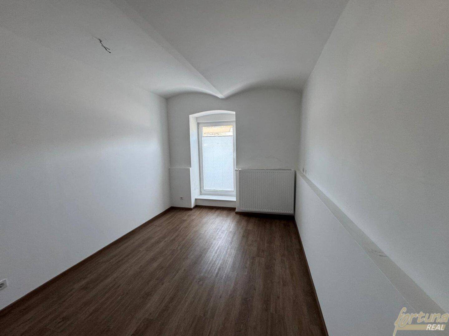 Heller Raum mit Laminatboden, weiß gestrichenen Wänden und einem kleinen Fenster mit Heizkörper.
