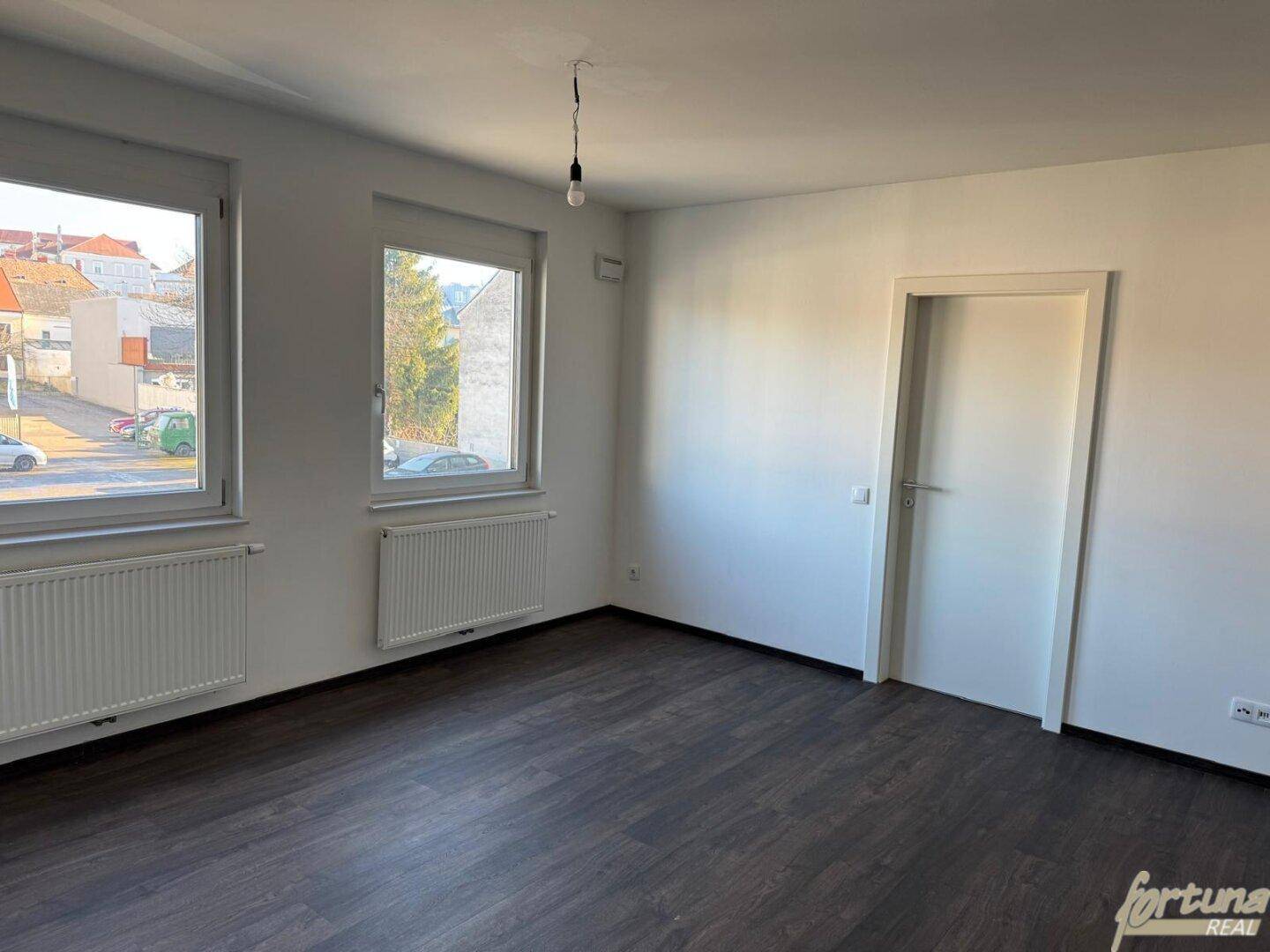Heller Raum mit zwei großen Fenstern, Laminatboden und weiß gestrichenen Wänden.