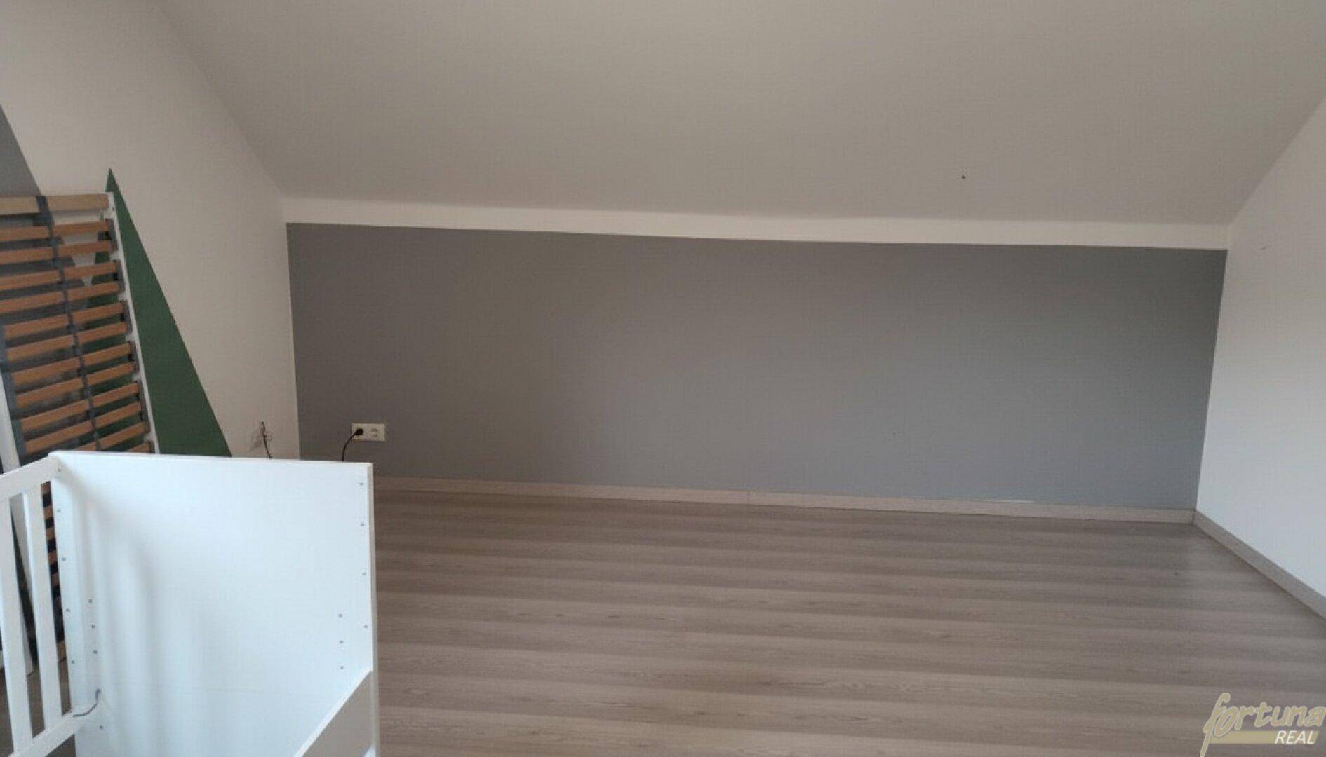 Leeres Zimmer mit schrägen Wänden, Holzboden und einer grauen Akzentwand.