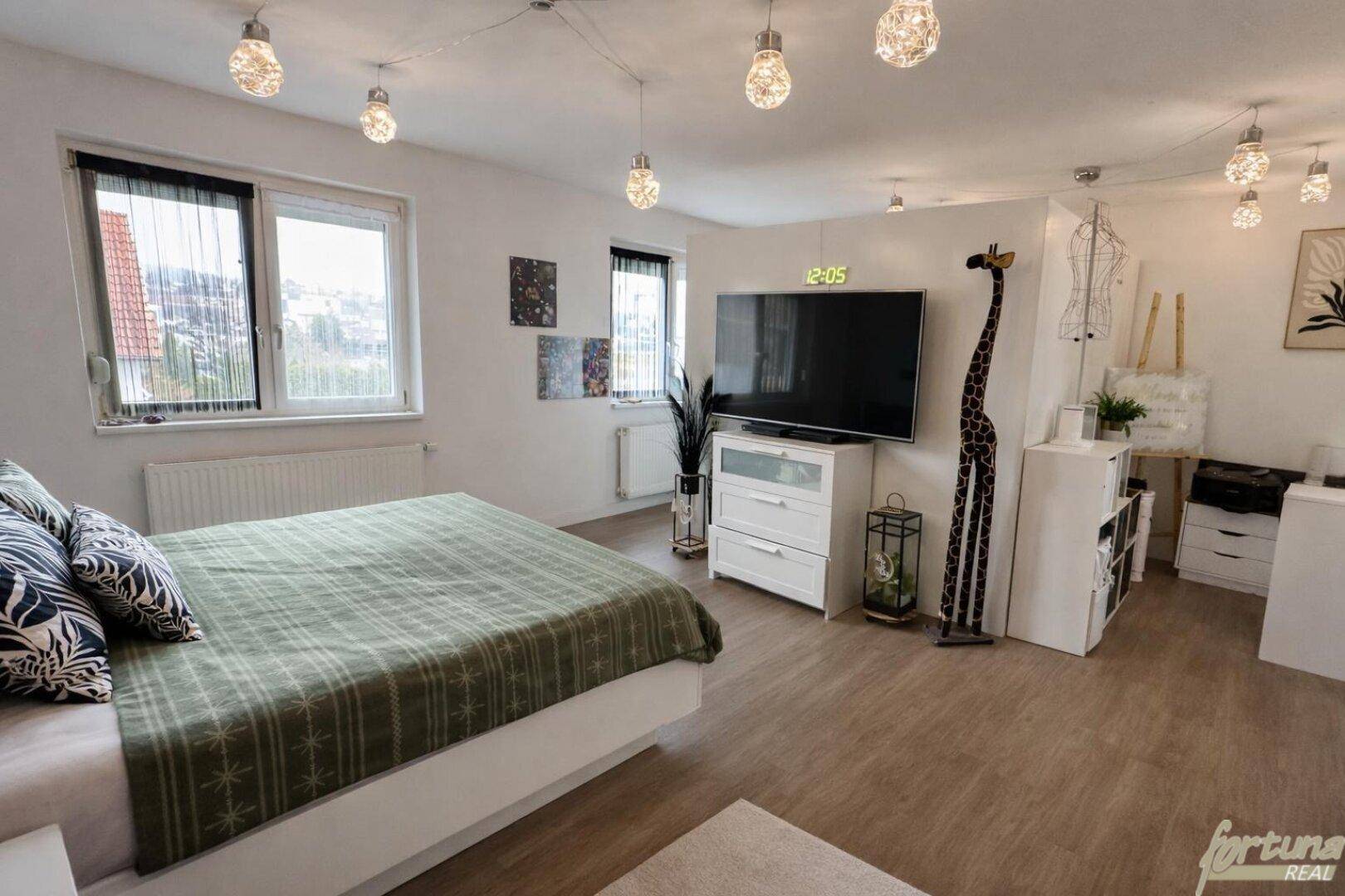 Helles Schlafzimmer mit großem Bett, Fernseher und dekorativer Beleuchtung.