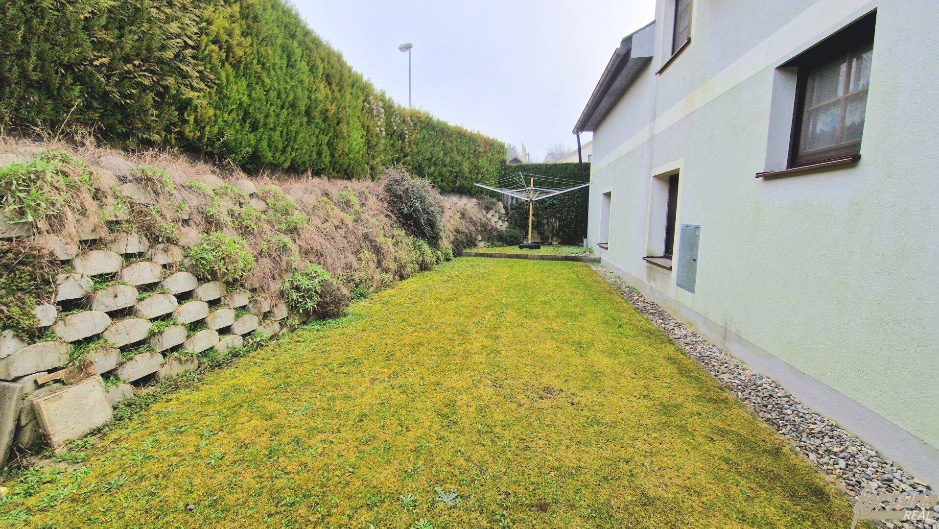 Schmaler Gartenstreifen entlang des Hauses mit Rasen und Stützmauer.