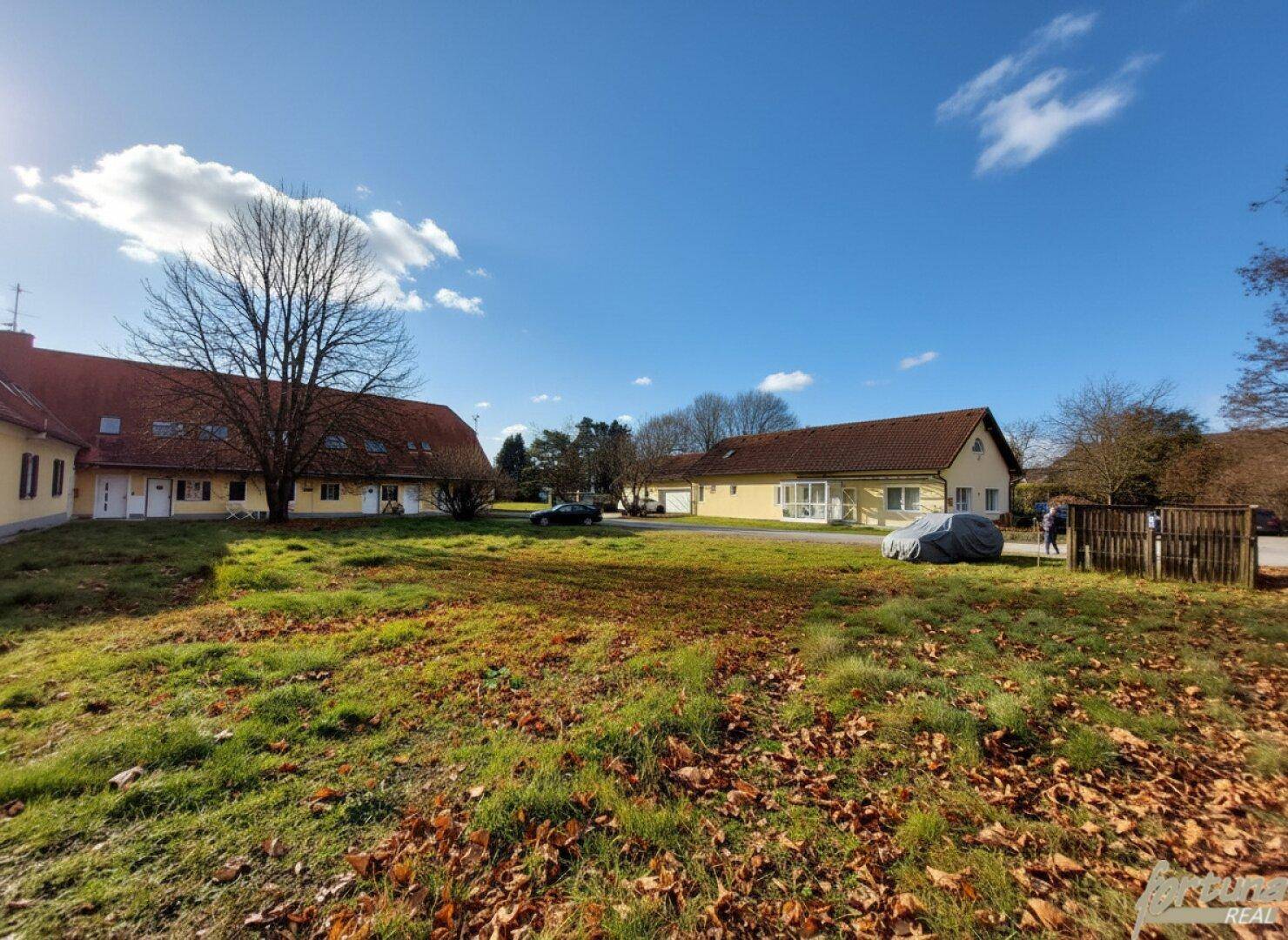 Weitläufige Grünfläche mit Herbstlaub und zwei gelben Gebäuden mit roten Dächern im Hintergrund unter blauem Himmel.