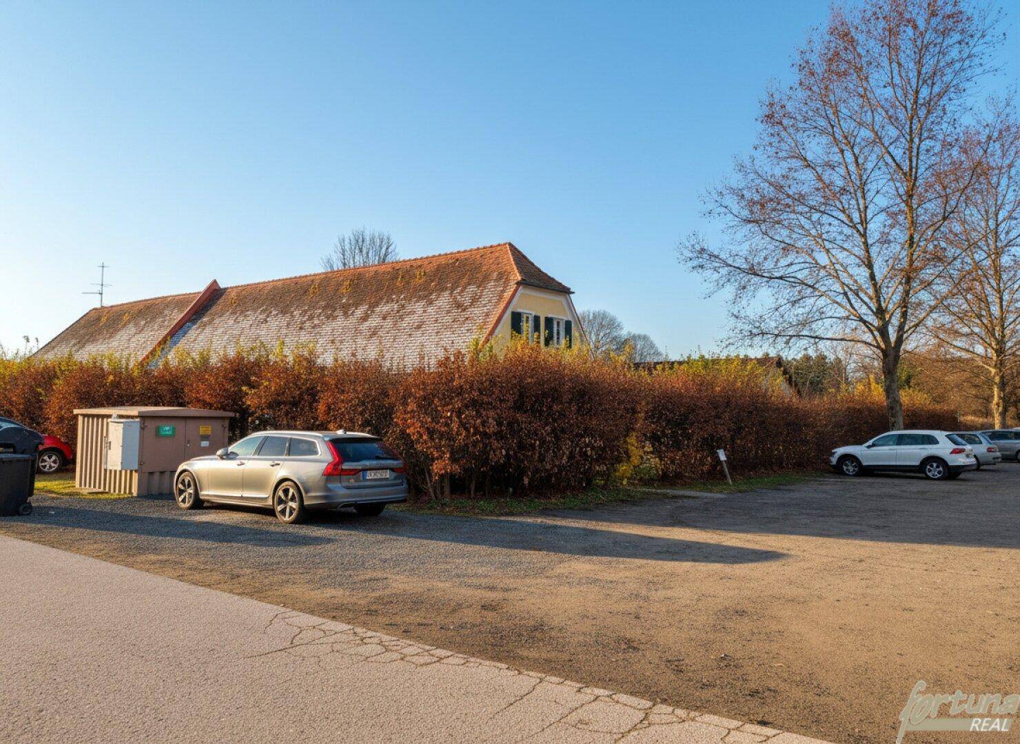 Parkplatz mit mehreren Fahrzeugen und einer Hecke, im Hintergrund ein gelbes Gebäude mit rotem Dach unter blauem Himmel.