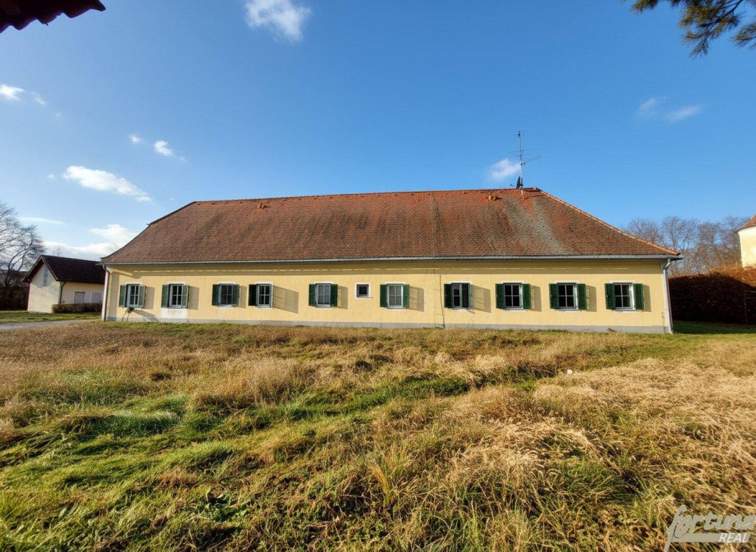 Langes, gelbes Gebäude mit rotem Dach und grünen Fensterläden, umgeben von einer sonnigen Grünfläche.