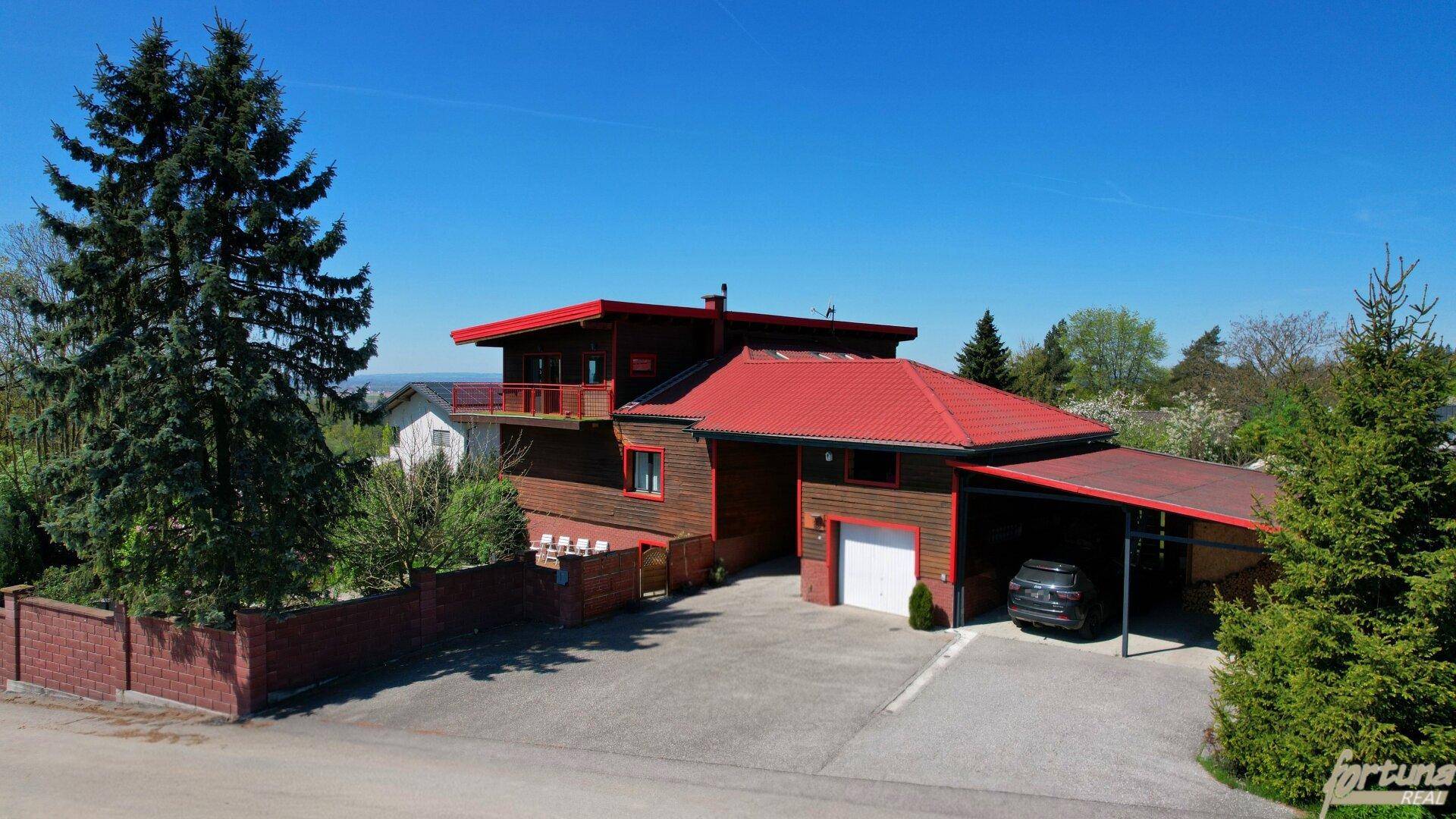 Großzügiges Holzhaus mit rotem Dach, Carport und weitläufiger Auffahrt in ländlicher Umgebung.