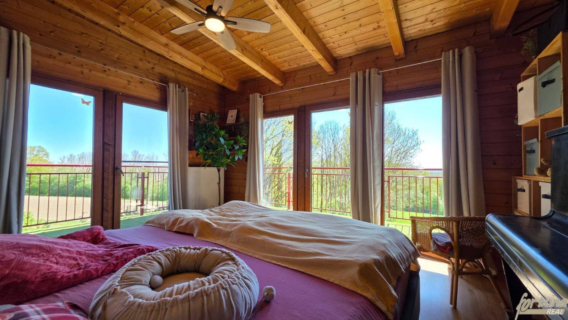 Gemütliches Schlafzimmer mit Holzbalkendecke, großen Fenstern und Zugang zum Balkon.