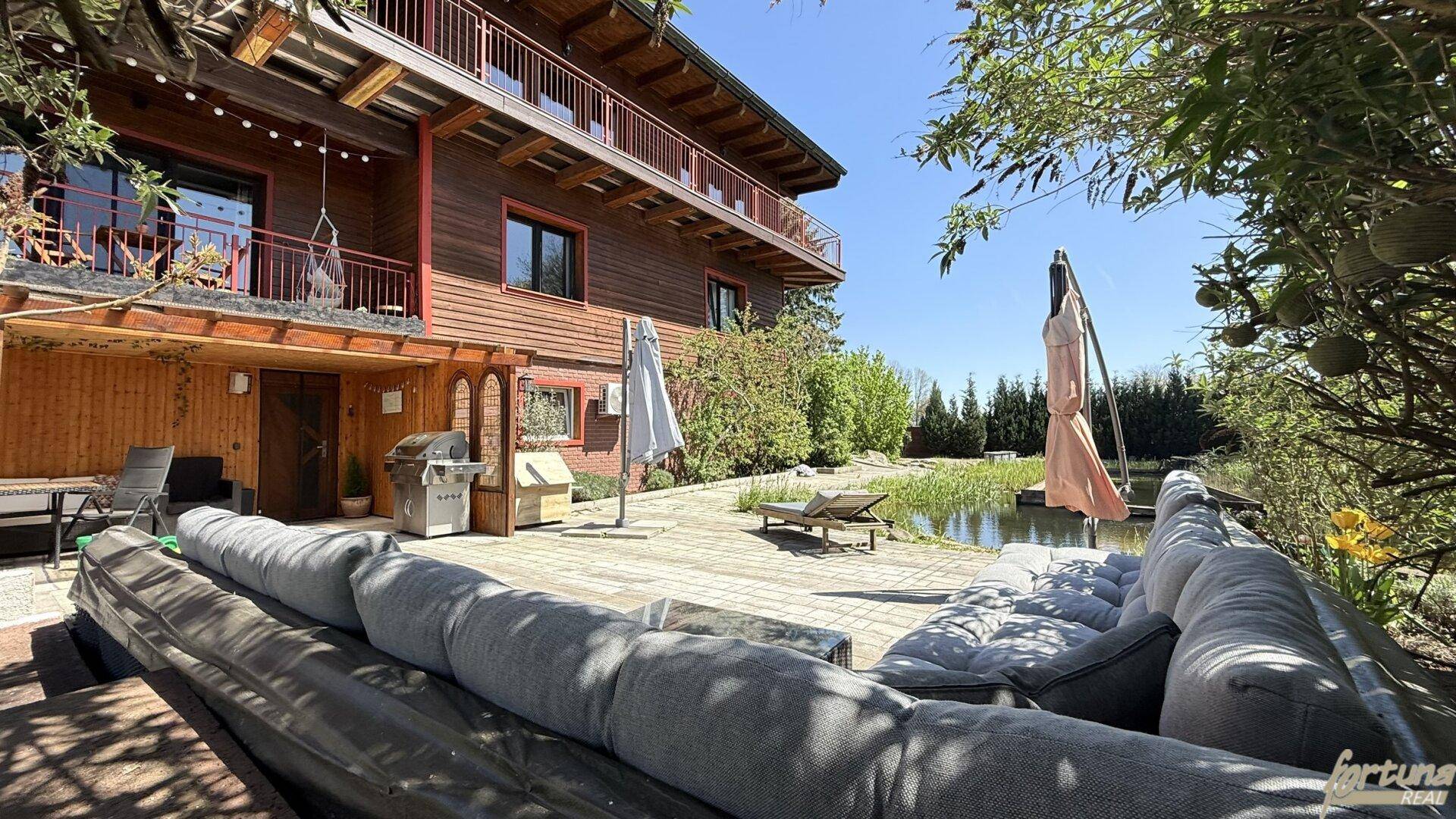 Holzhaus mit großer Terrasse, Outdoor-Sitzbereich und einem natürlichen Teich im Garten.