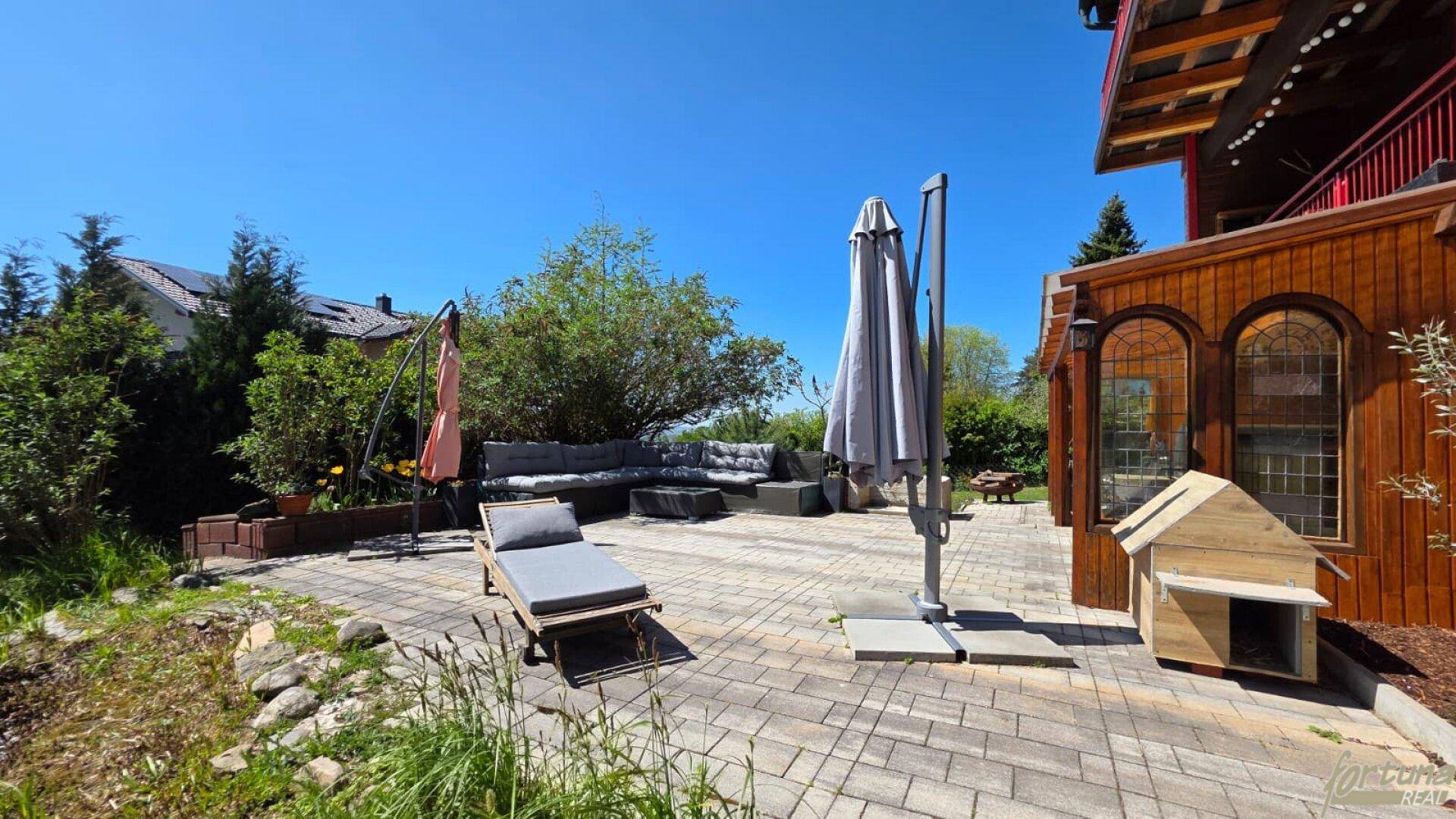 Sonniger Außenbereich mit gepflasterter Terrasse, Liegestuhl und Hundehütte.