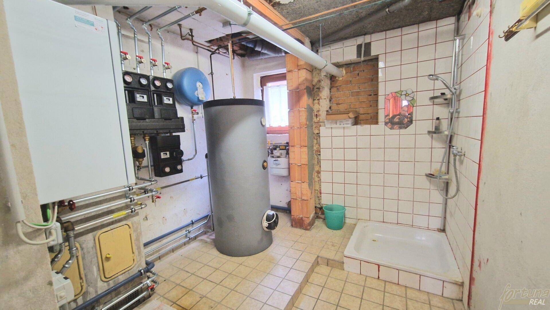Heizungsraum im Keller mit Warmwasserspeicher und einfacher Duschkabine, funktional eingerichtet.