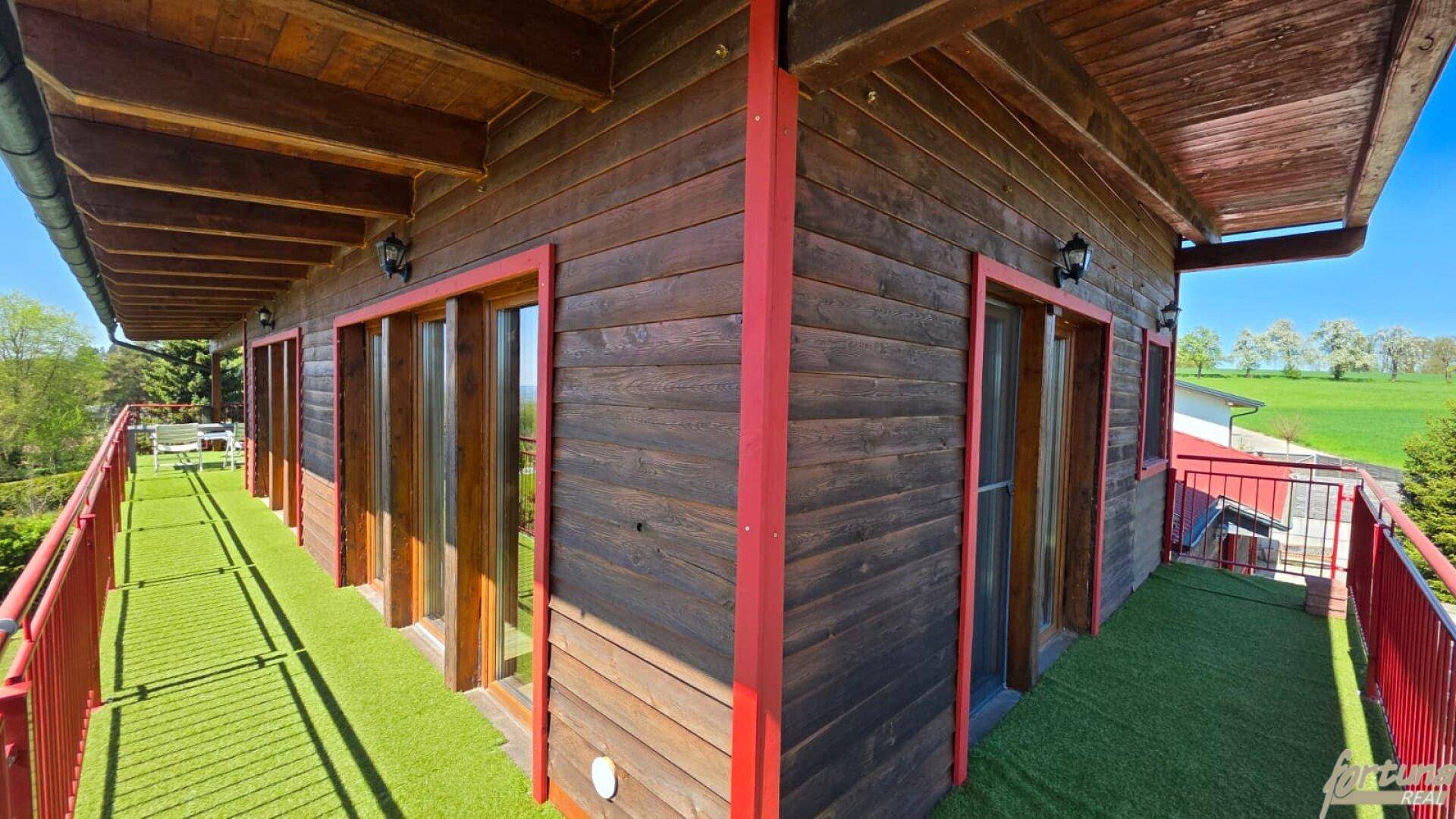 Langer Balkon mit Holzwänden, rotem Geländer und Kunstrasen, umgeben von grüner Landschaft.