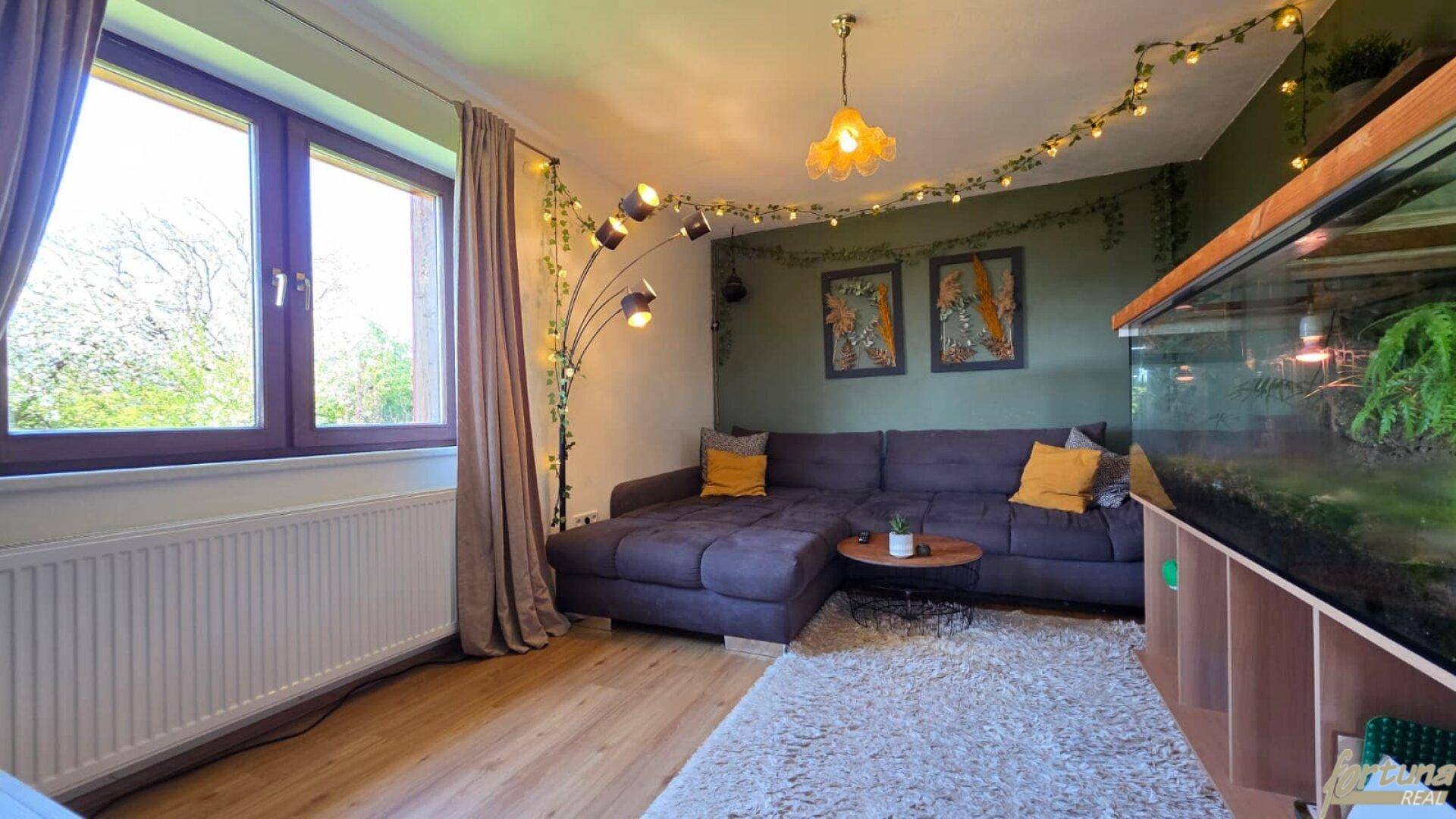 Gemütliches Wohnzimmer mit großem Sofa, Couchtisch, Aquarium und viel Tageslicht durch das Fenster.