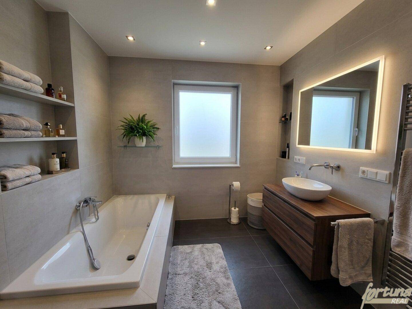 Modernes Badezimmer mit Badewanne, Waschtisch und beleuchtetem Spiegel in neutralen Grautönen.