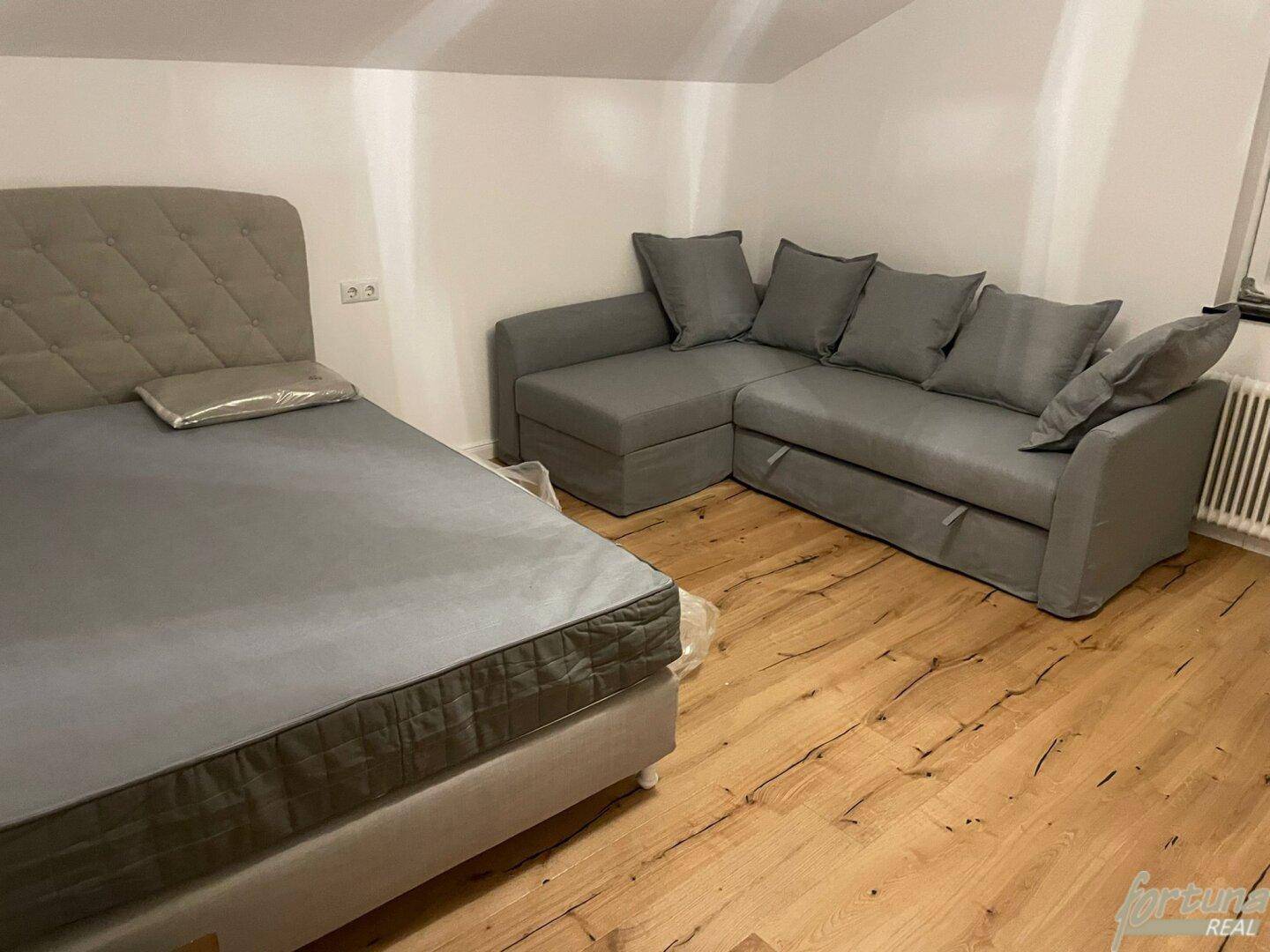 Schlafbereich im Dachgeschoss mit Bett, grauem Sofa und Holzfußboden.