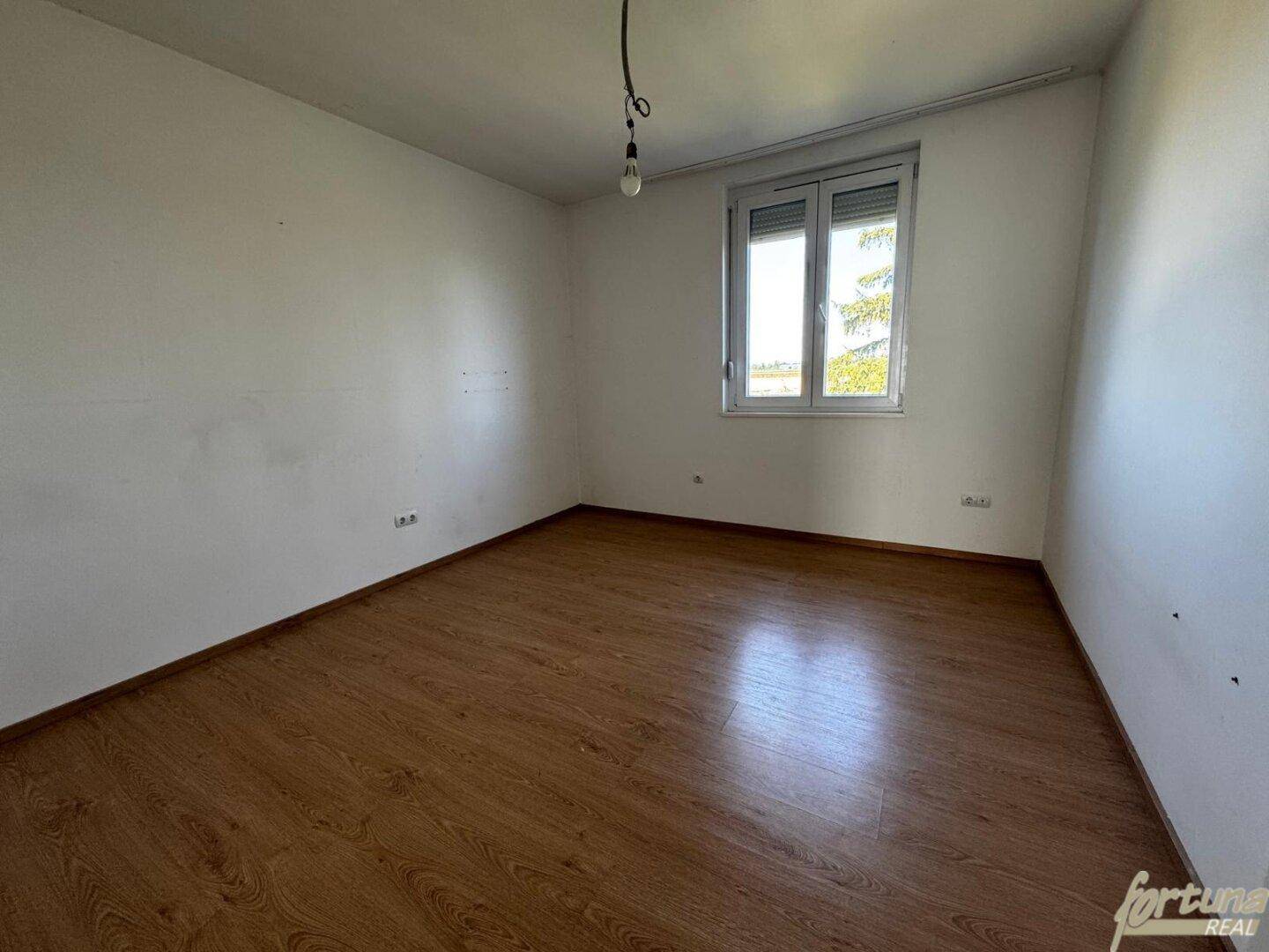 Helles Schlafzimmer mit Laminatboden und einem Fenster mit Blick ins Grüne.