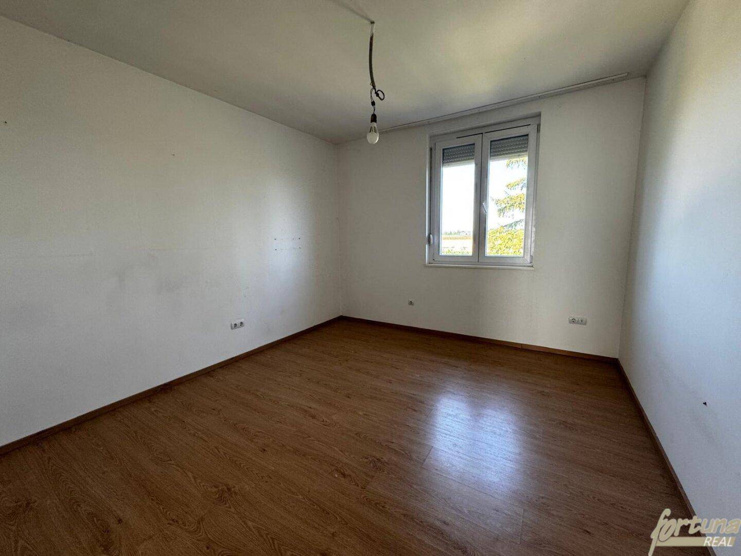 Helles Schlafzimmer mit Laminatboden und einem Fenster mit Blick ins Grüne.