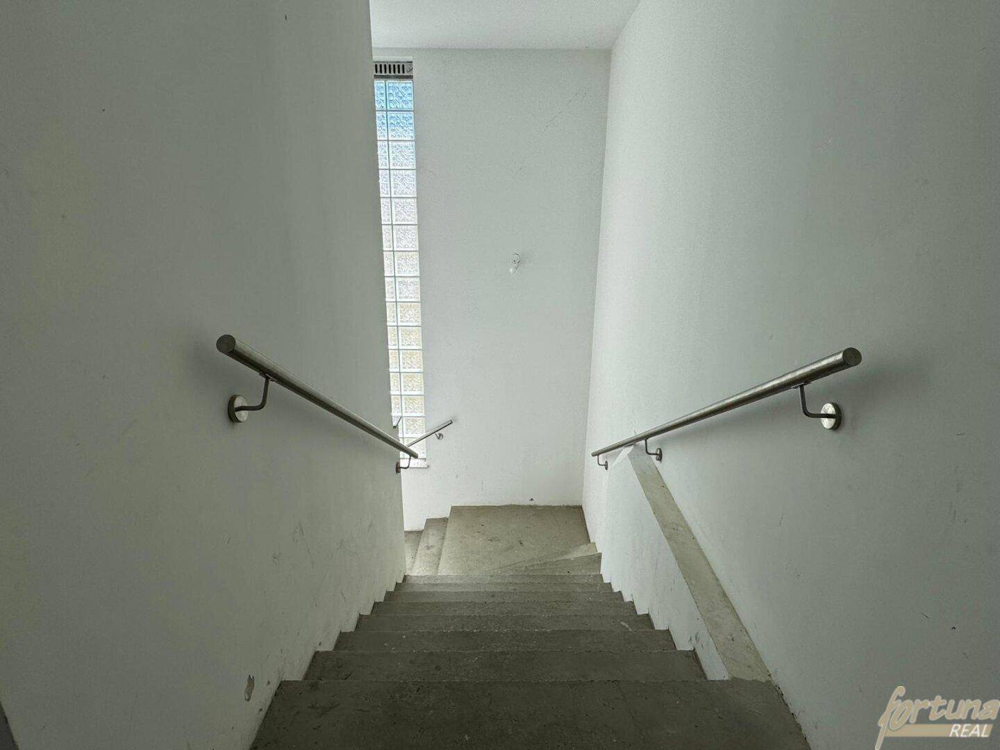 Treppenhaus mit Betonstufen und Handläufen, beleuchtet durch vertikale Glasbausteine.