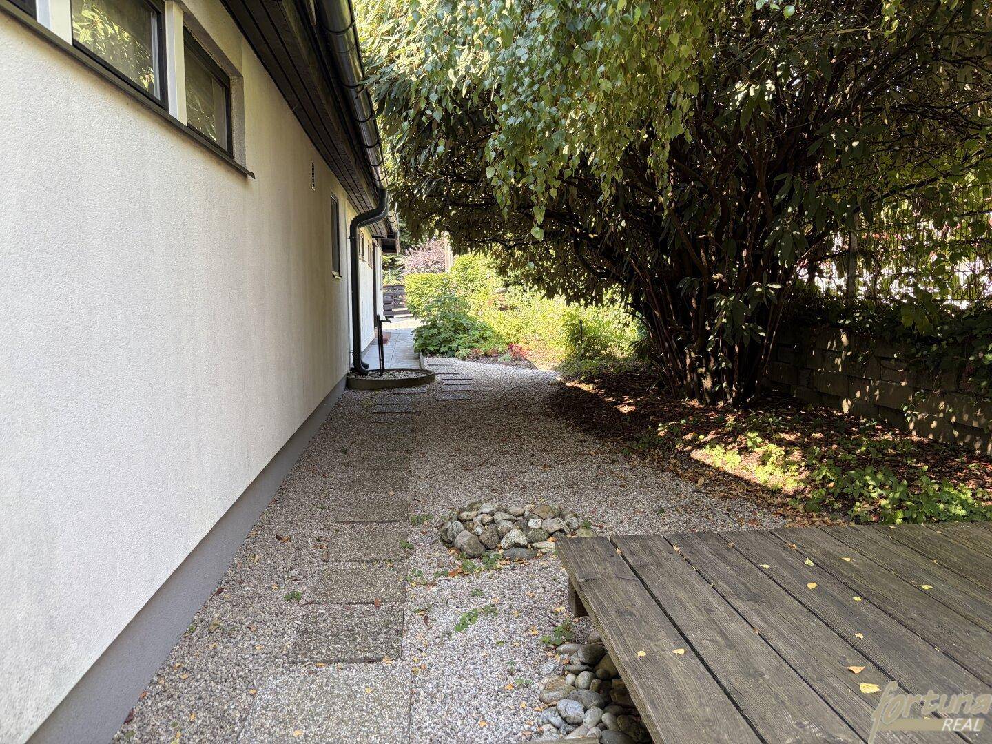 Seitenweg entlang des Hauses mit Kies, Trittsteinen und üppiger Bepflanzung.