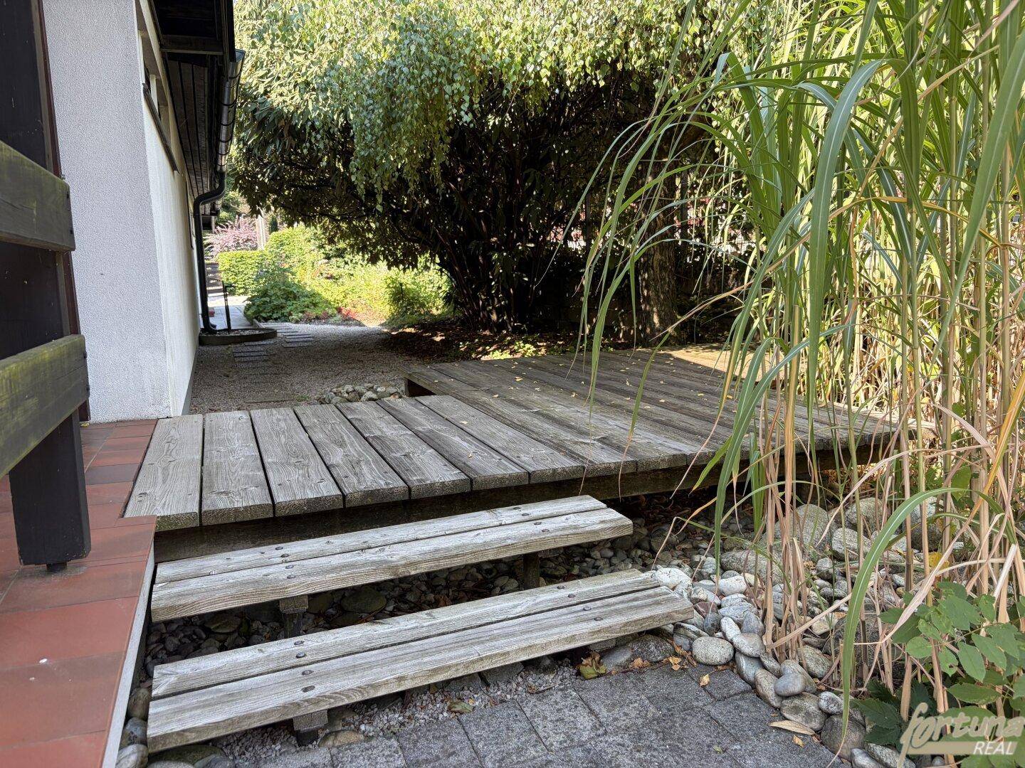 Holzsteg und Stufen führen durch den Gartenbereich mit Kies und hohen Gräsern.