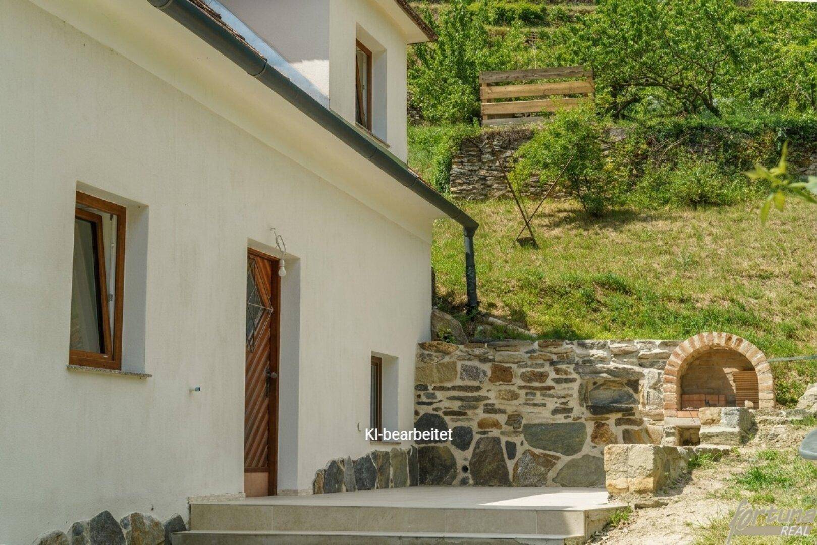 Seitlicher Blick auf das Haus mit einer kleinen Terrasse und einem traditionellen Steinofen im Freien.