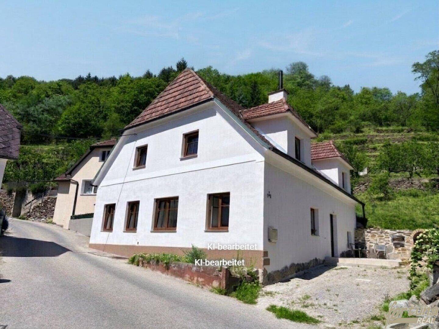 Charmantes weißes Haus mit Ziegeldach an einer Hanglage, umgeben von grüner Natur.