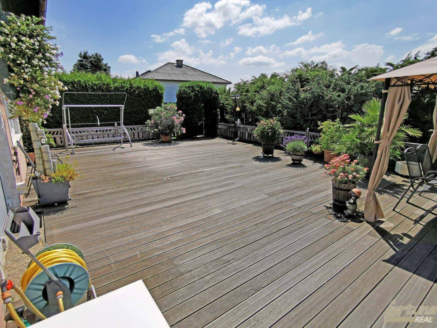 Große Holzterrasse mit Schaukel, Pflanzen und Zugang zum Gartenbereich.