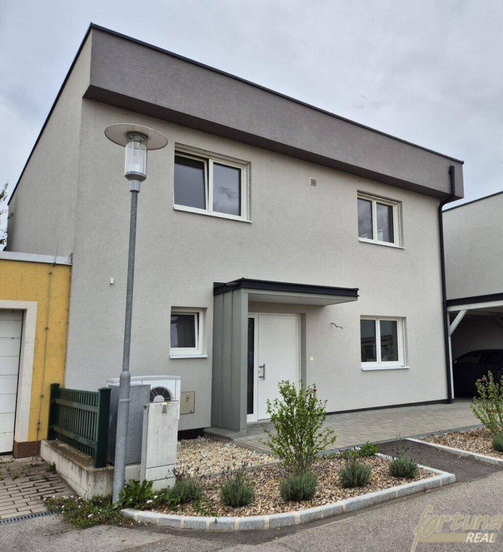 Moderne Hausfassade mit Eingangsbereich, kleinem Vorgarten und Carport im Hintergrund.