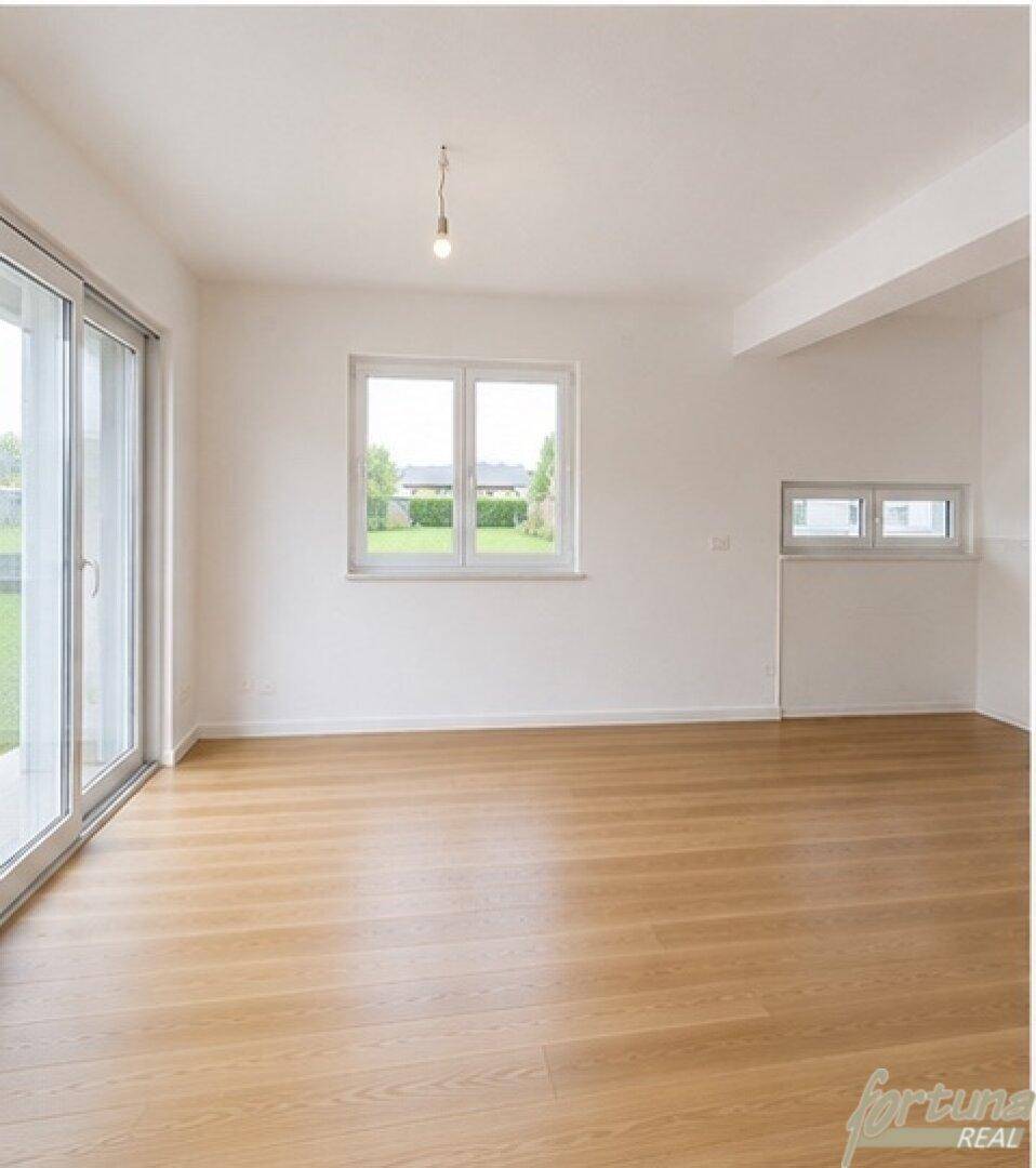 Geräumiges Wohnzimmer mit hellem Holzfußboden, weißen Wänden und mehreren Fenstern.